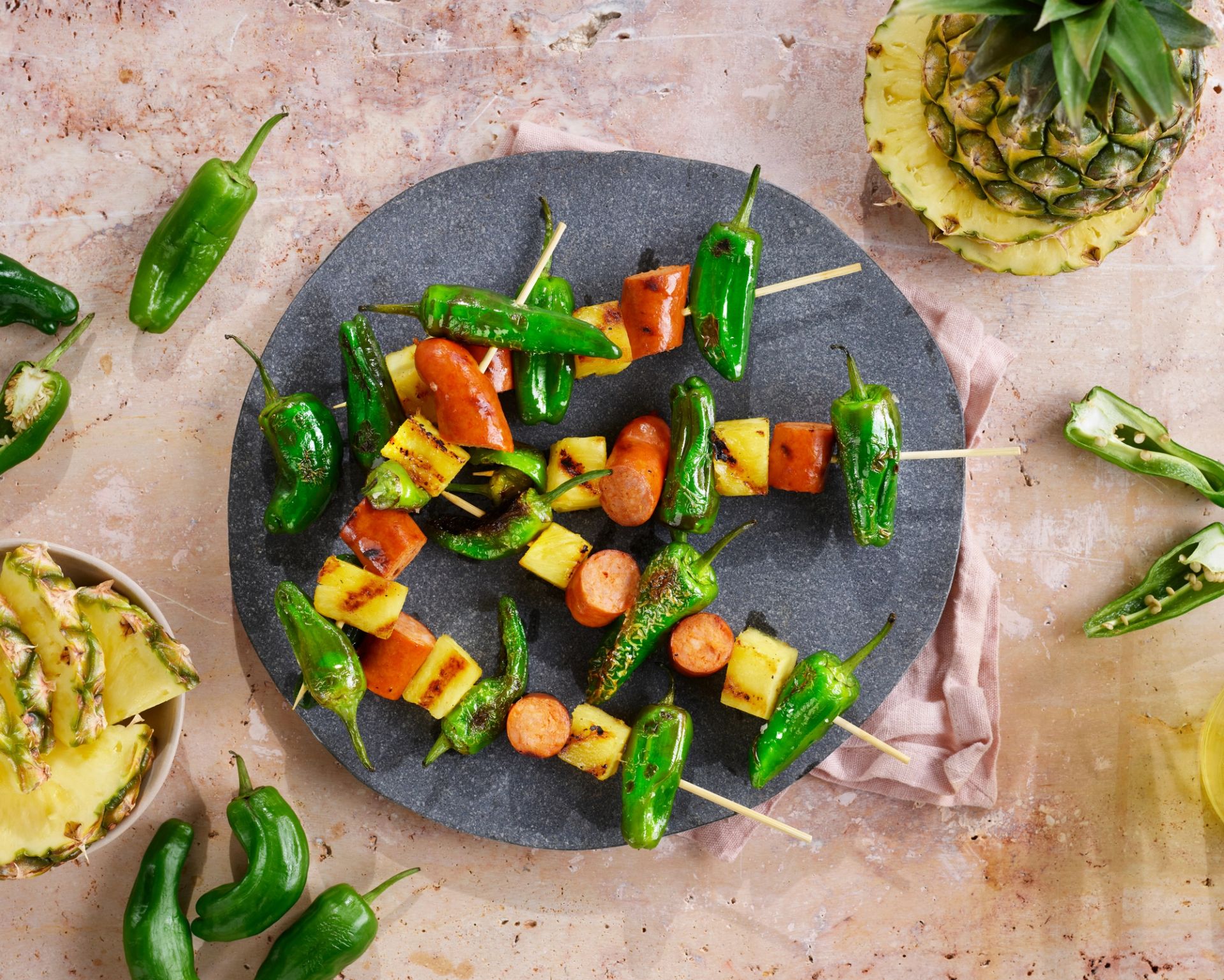 Grillspyd med Padron-paprika, chorizo og grillet ananas servert på en rund, grå steinfjøl. Rundt fjølen ligger rå Padrón-paprika, en fersk ananas og en bolle med ananasbiter.