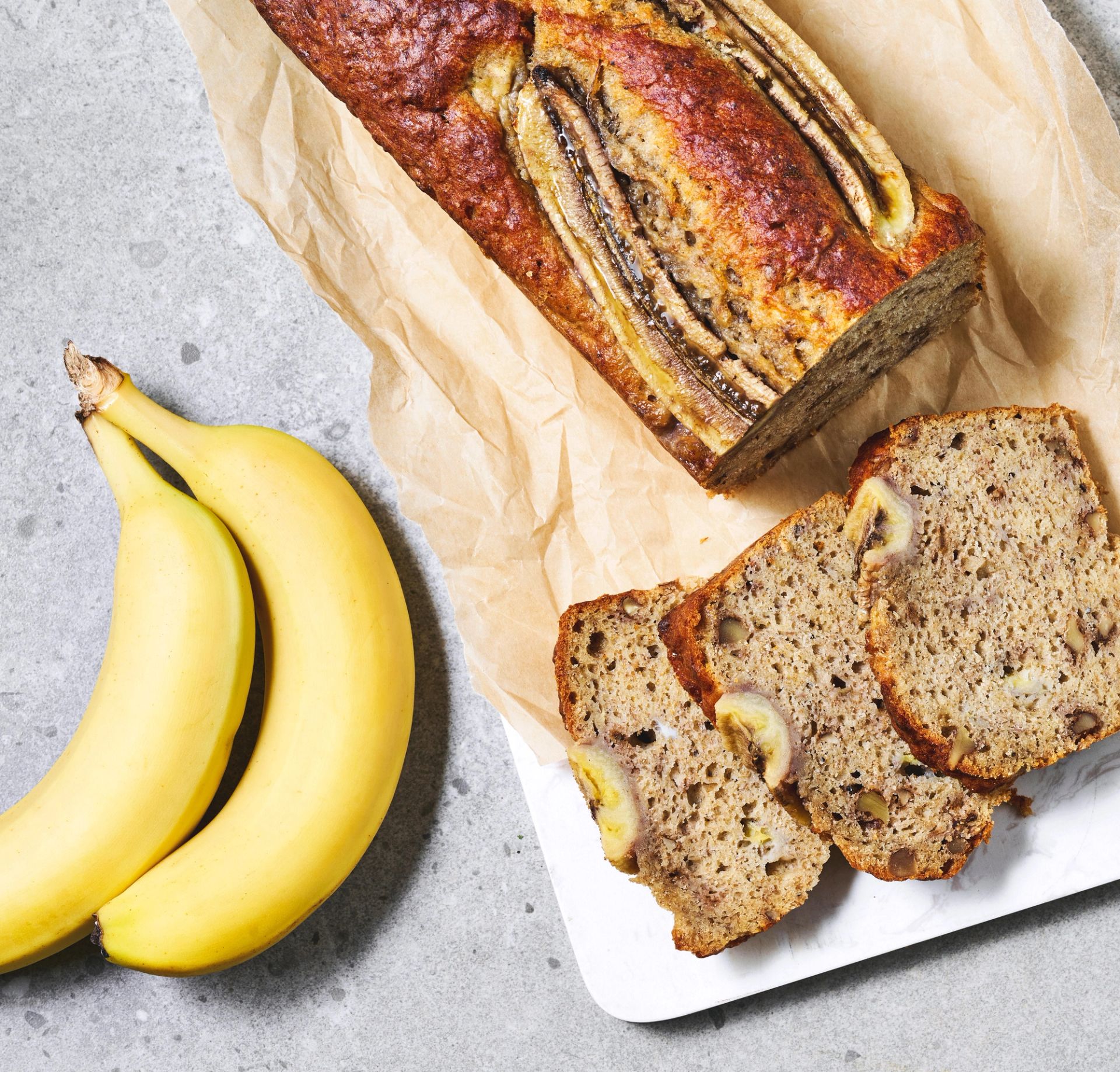 Saftig bananbrød med synlige bananskiver på toppen ligger på bakepapir ved siden av flere oppskårne skiver. To hele bananer er plassert ved siden av brødet, og den myke konsistensen med biter av banan kommer tydelig frem i skivene.