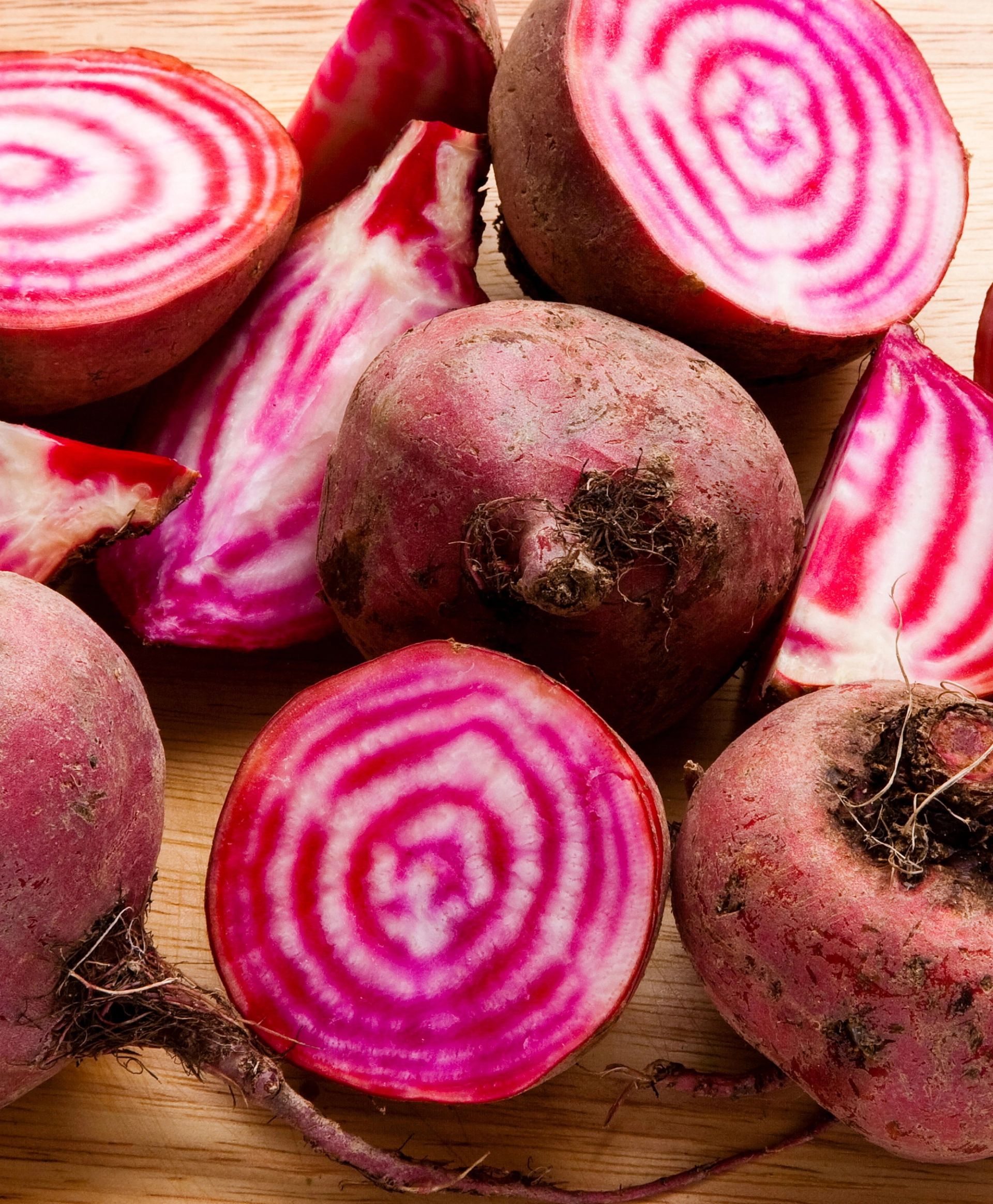 Rødbeter av typen Chioggia, både hele og kuttet i skiver og biter, lagt på et trefarget underlag. De kuttede rødbetene viser tydelige rosa og hvite konsentriske ringer.