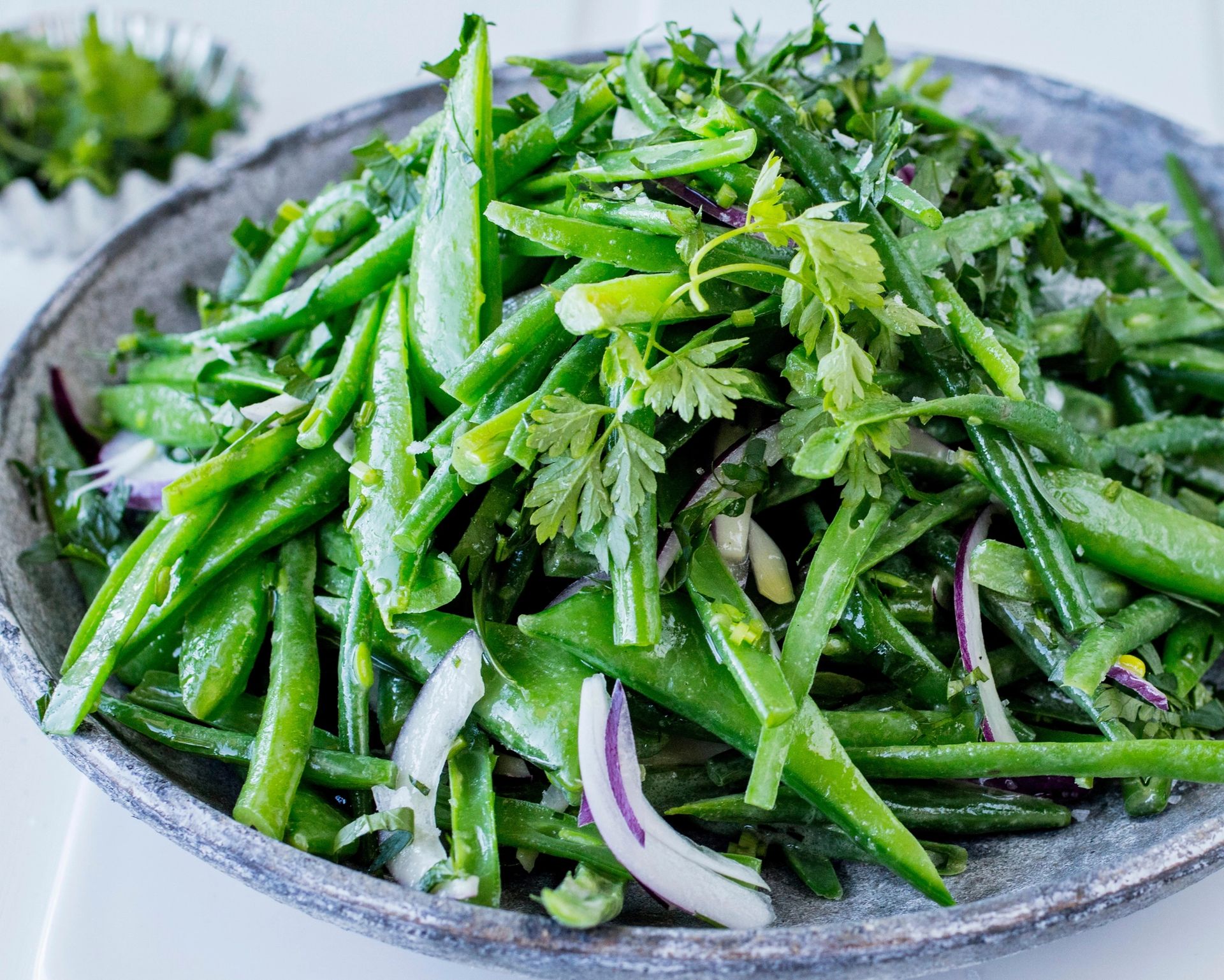 En stor bolle fylt med en grønn bønnesalat med sukkererter, skiver av rødløk og friske urter, lett glinsende av dressing, servert på en lys overflate med en nøytral bakgrunn.