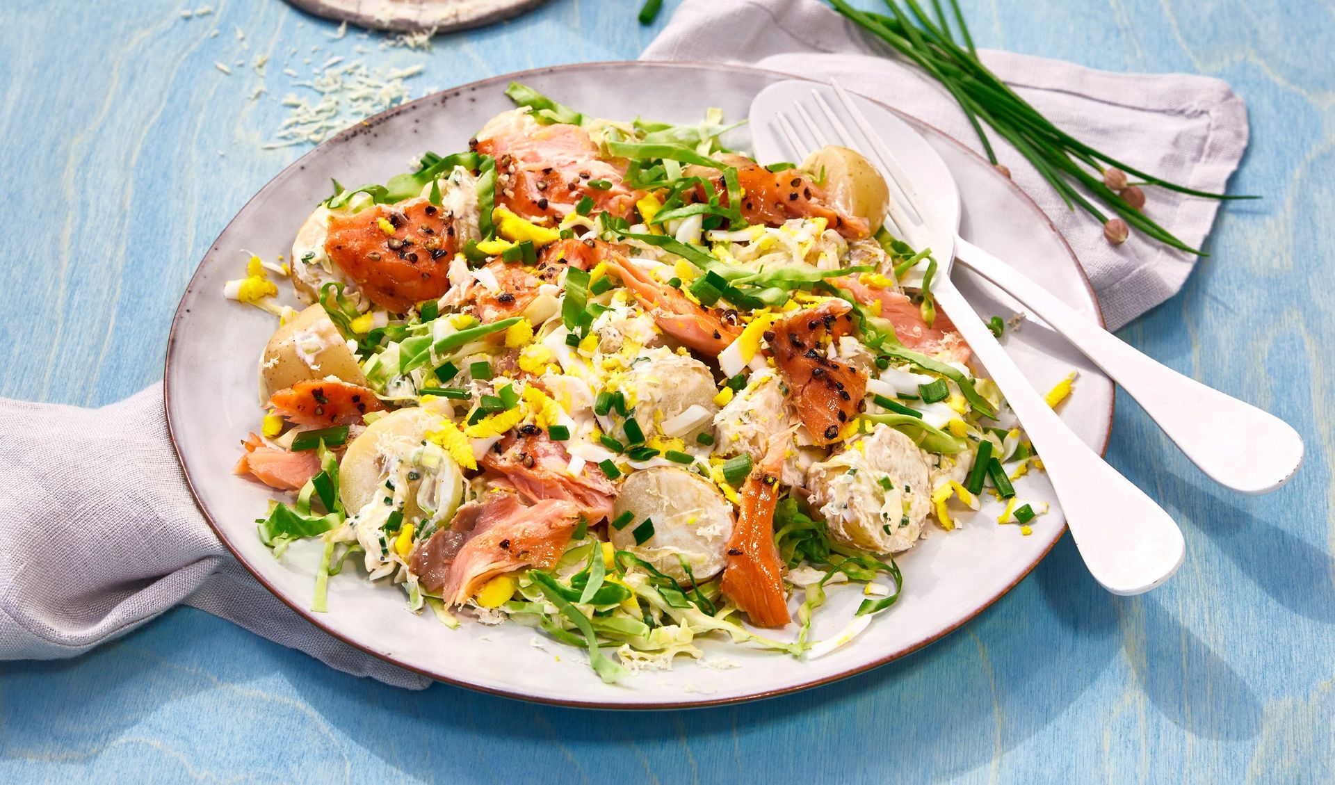 Salat med røkt laks, finsnittet kål, kokte poteter og hakket gressløk, toppet med sesamfrø og servert på et stort fat på et lyseblått bord.