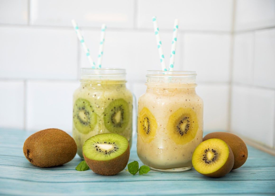 Smoothie med kiwi servert i glasskrus med sugerør, dekorert med skiver av grønn og gul kiwi, med fersk kiwi på bordet.