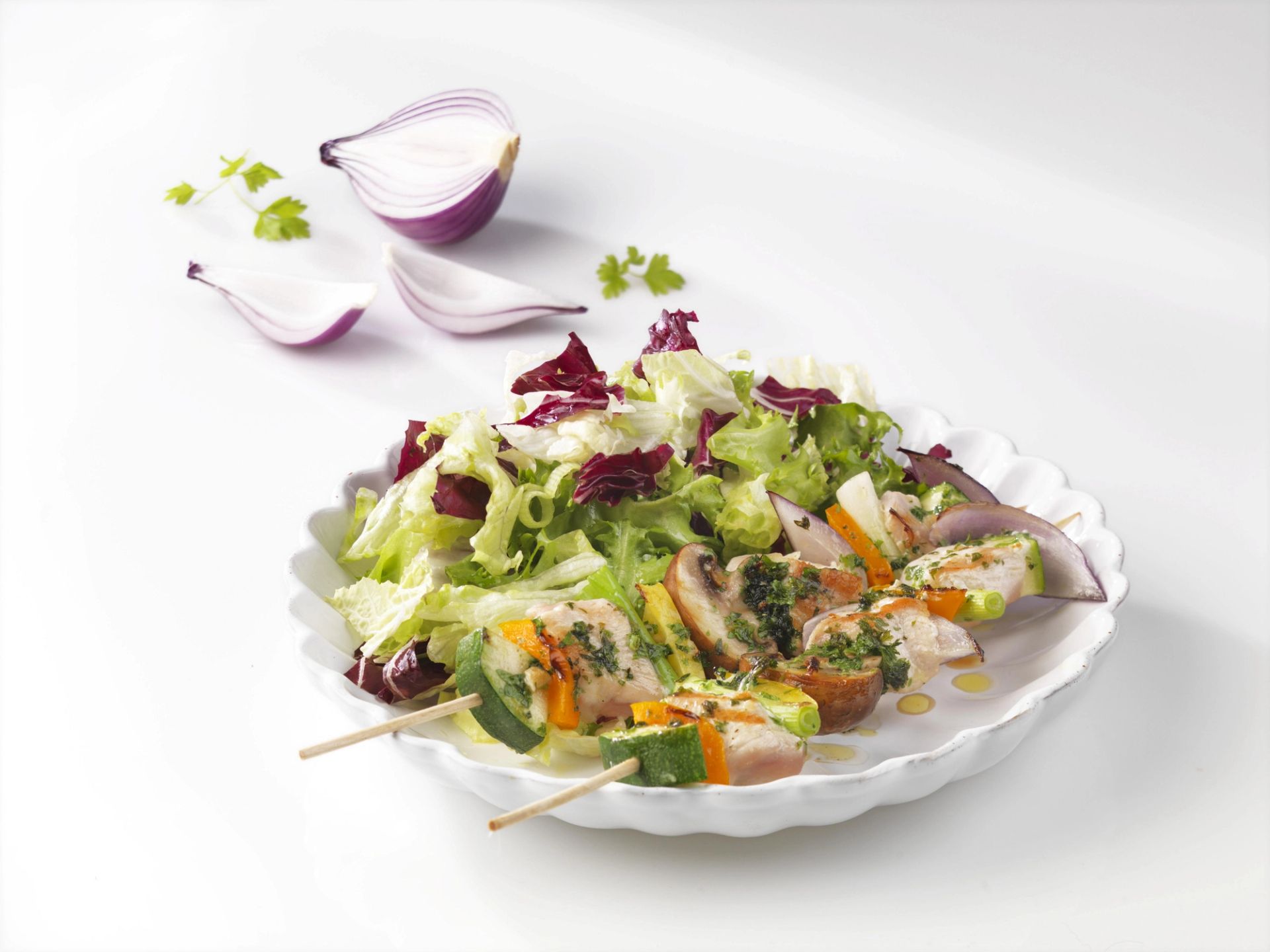 Salattallerken med blandede salatblader, grillet grønnsaksspyd og skiver av rødløk, servert på hvit tallerken.