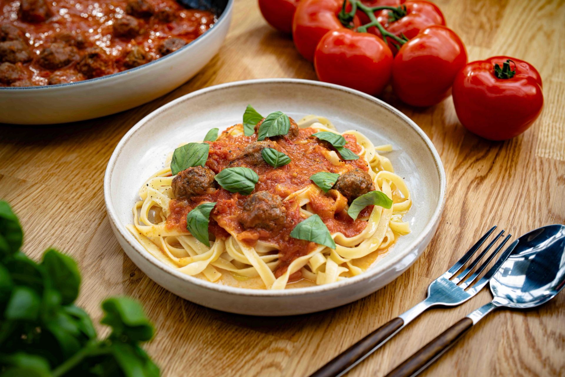 Tallerken med pasta og tomatsaus toppet med kjøttboller og frisk basilikum, servert på et trebord med tomater i bakgrunnen.