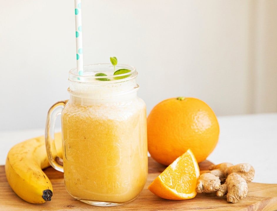 Smoothie med ingefær, appelsin og banan servert i glasskrus med sugerør, pyntet med mynte, med fersk appelsin, banan og ingefær ved siden av.