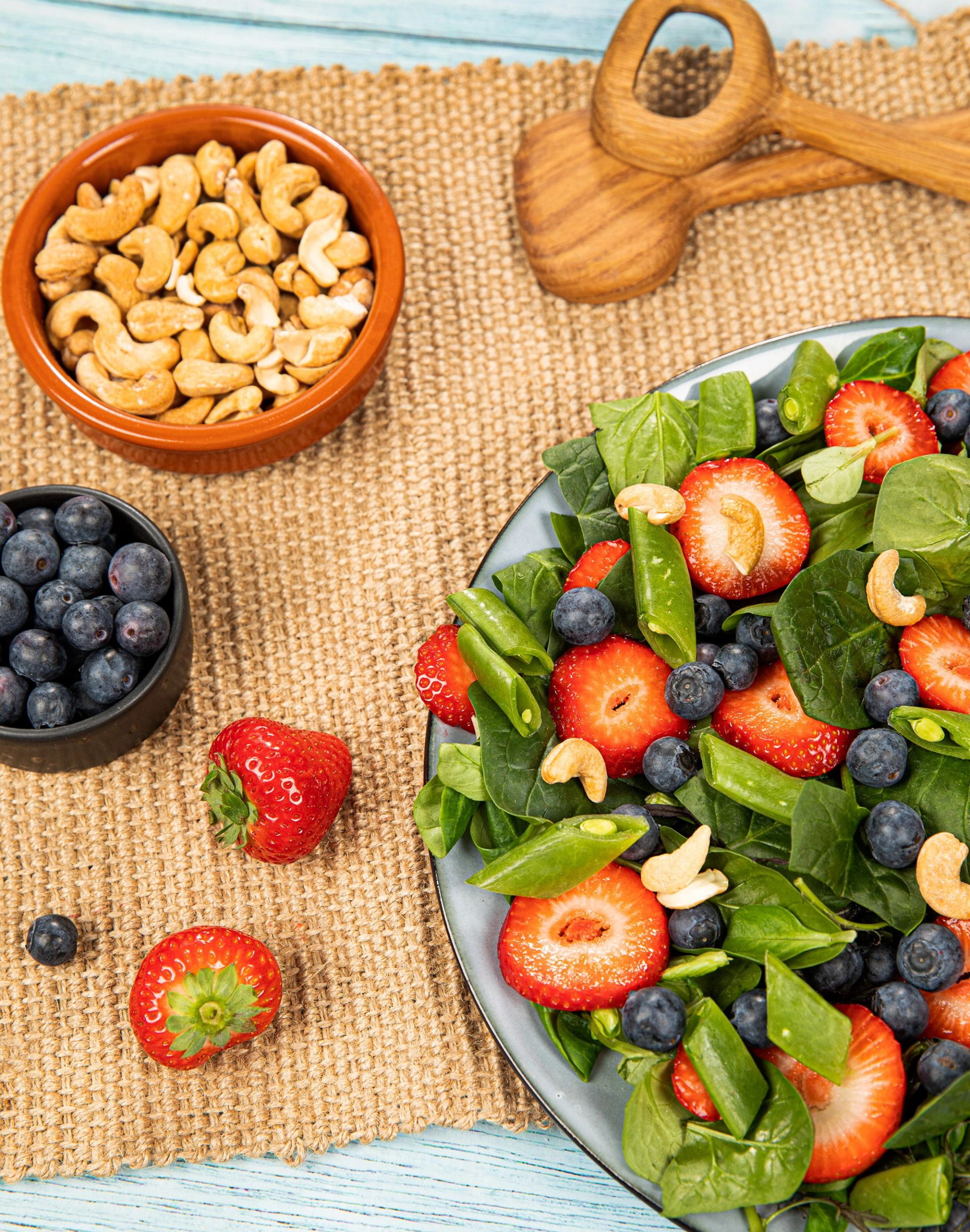 En tallerken med salat laget av frisk spinat, halve jordbær, blåbær og cashewnøtter står på en overflate dekket med et juteliknende underlag. Ved siden av står en liten skål med blåbær, en terrakottaskål med cashewnøtter og et treskje‑redskap. Flere jordbær og noen løse blåbær ligger spredt rundt skålene.