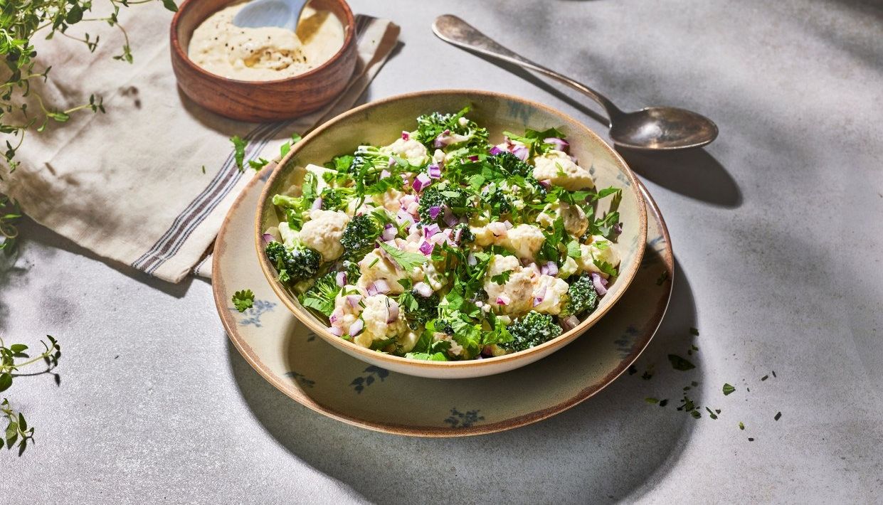 Blomkål‑ og brokkolisalat med rødløk og frisk bladpersille servert i en bolle, med kremet dressing i en separat skål og grønne urter spredt rundt på et lyst bord.