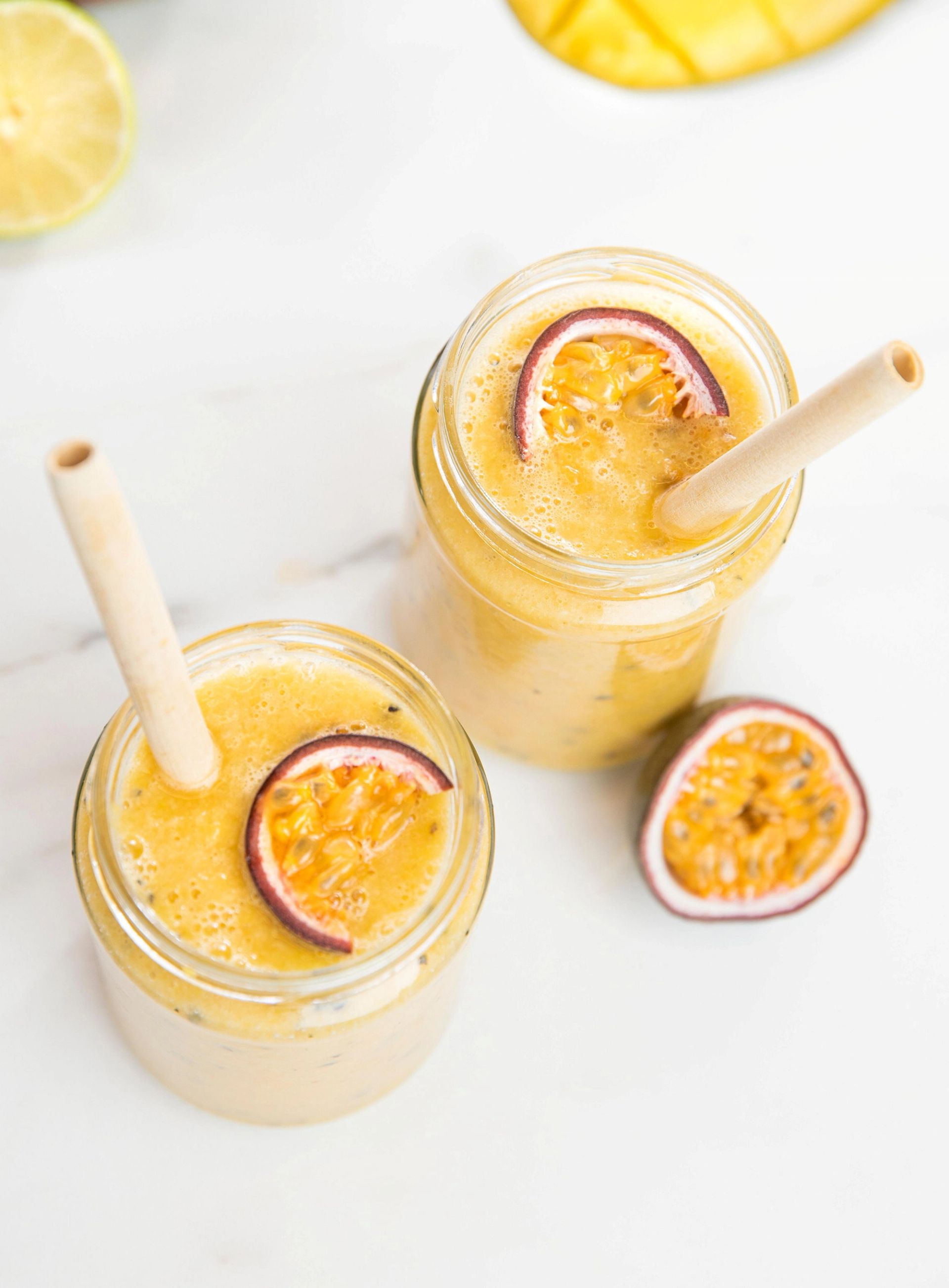 To glass med lys oransje smoothie toppet med skiver av pasjonsfrukt, servert med sugerør på en lys overflate, med sitrusfrukt og oppskåret mango i bakgrunnen.