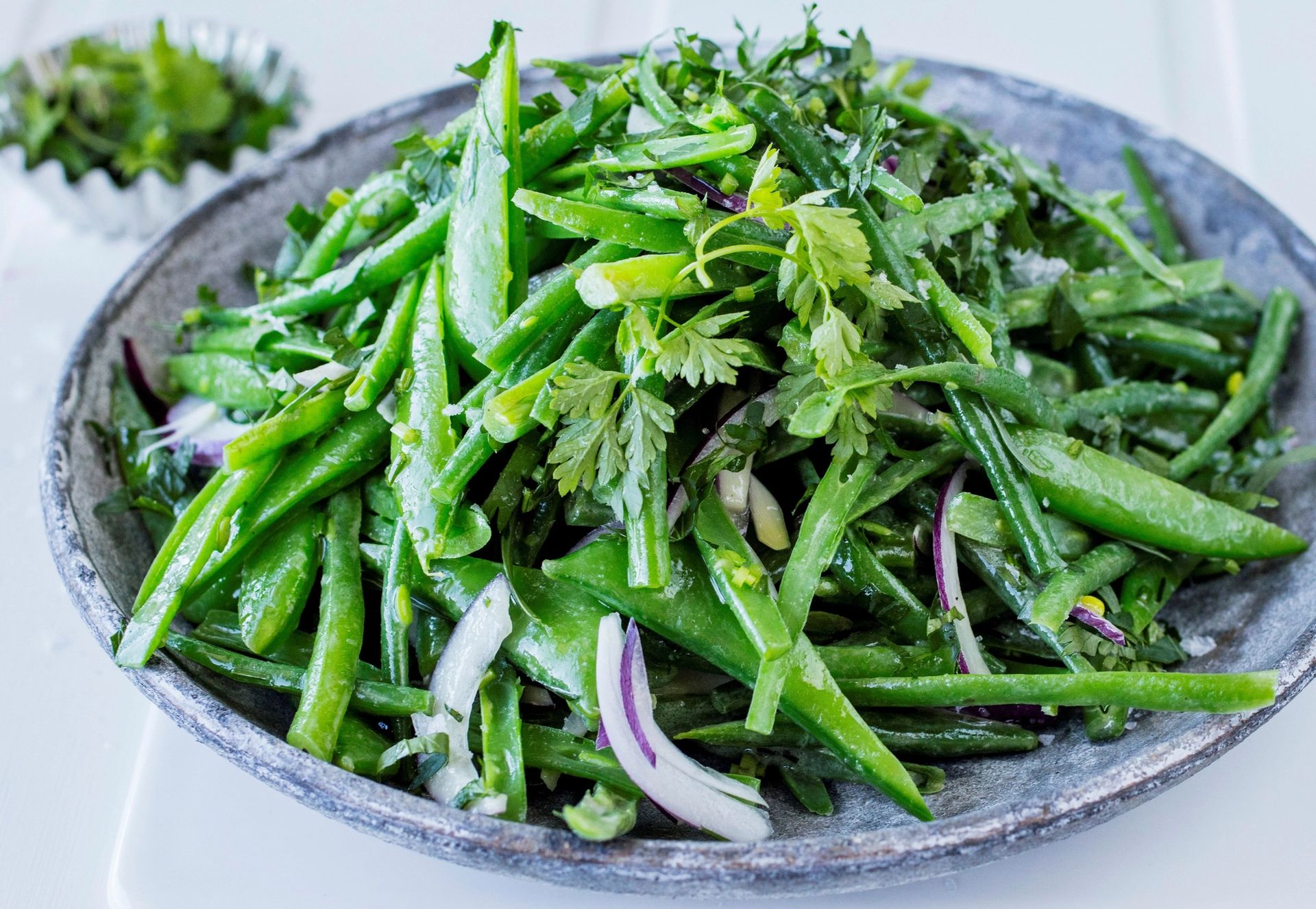 n frisk salat i en grå keramikkbolle med sprø grønne bønner, sukkererter og tynne skiver rødløk, toppet med hakkede urter og vendt i en lett dressing som gir en glinsende overflate.