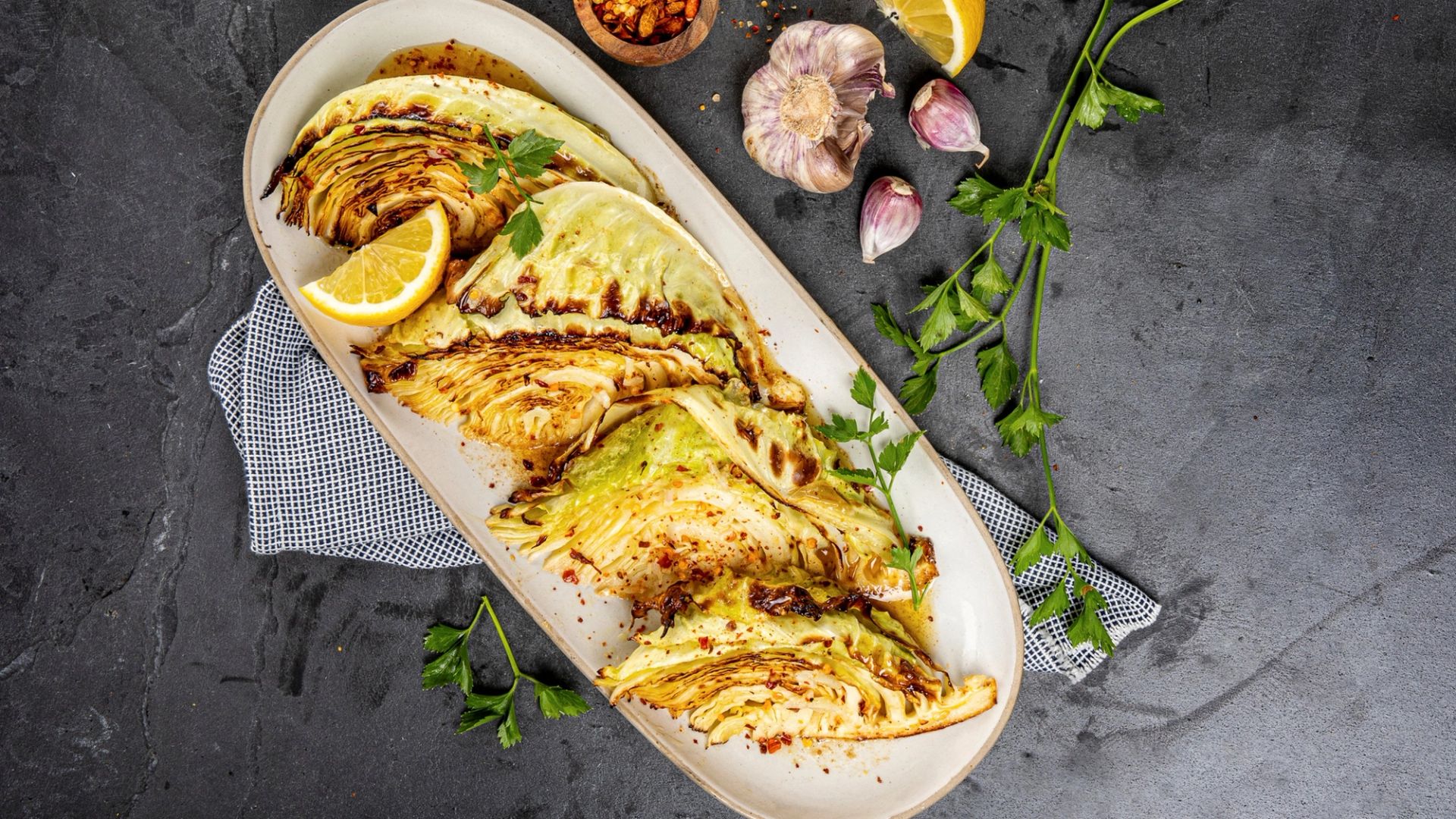 Grillede kålkløfter servert på et avlangt fat, krydret med chiliflak og garnert med sitronbåter, hvitløk og fersk persille på en mørk bakgrunn.