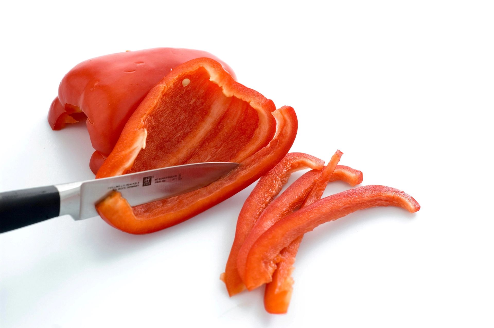 Rød paprika som skjæres i strimler med kniv på hvit bakgrunn.