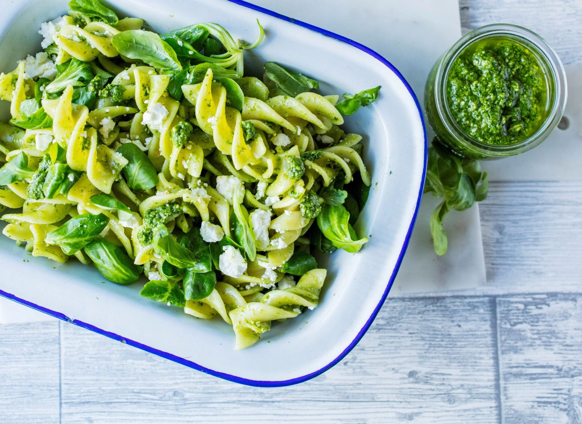 Pastasalat med fusilli, pesto, små biter av feta og friske basilikumblader i et hvitt fat med blå kant, og et glass med fersk, grønn pesto ved siden av.