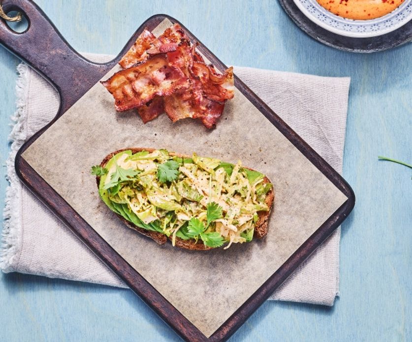 Ristet brødskive med avokado og salat toppet med urter, servert på brett med sprøstekt bacon ved siden av.