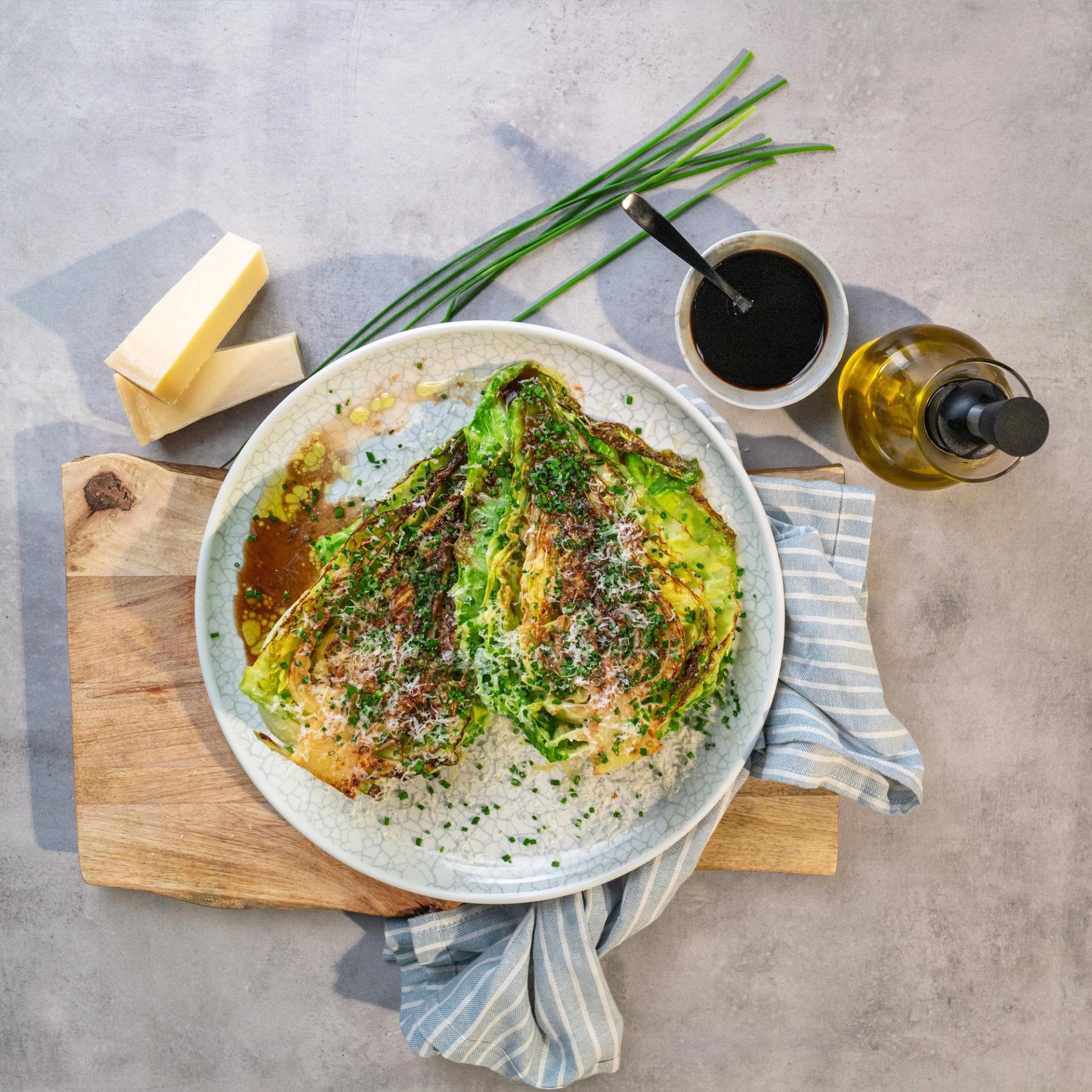Grillet spisskål servert på en tallerken, toppet med urter og revet ost, omgitt av smør, gressløk, dressing og olivenolje på et skjærebrett