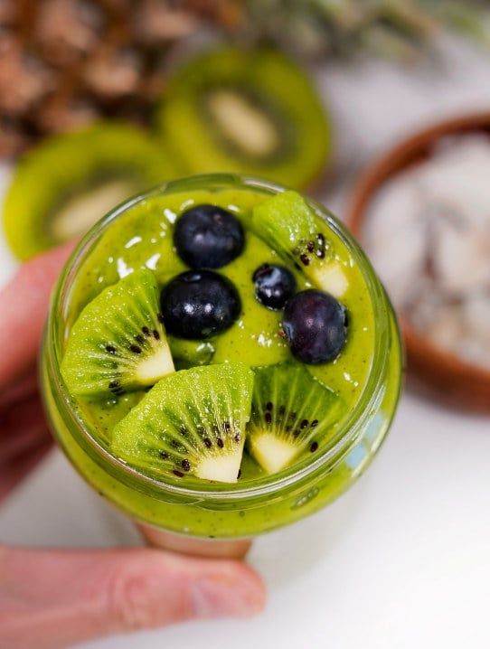 Grønn smoothie i et glass toppet med kiwiskiver og blåbær, holdt med venstre hånd, med kiwi i bakgrunnen.