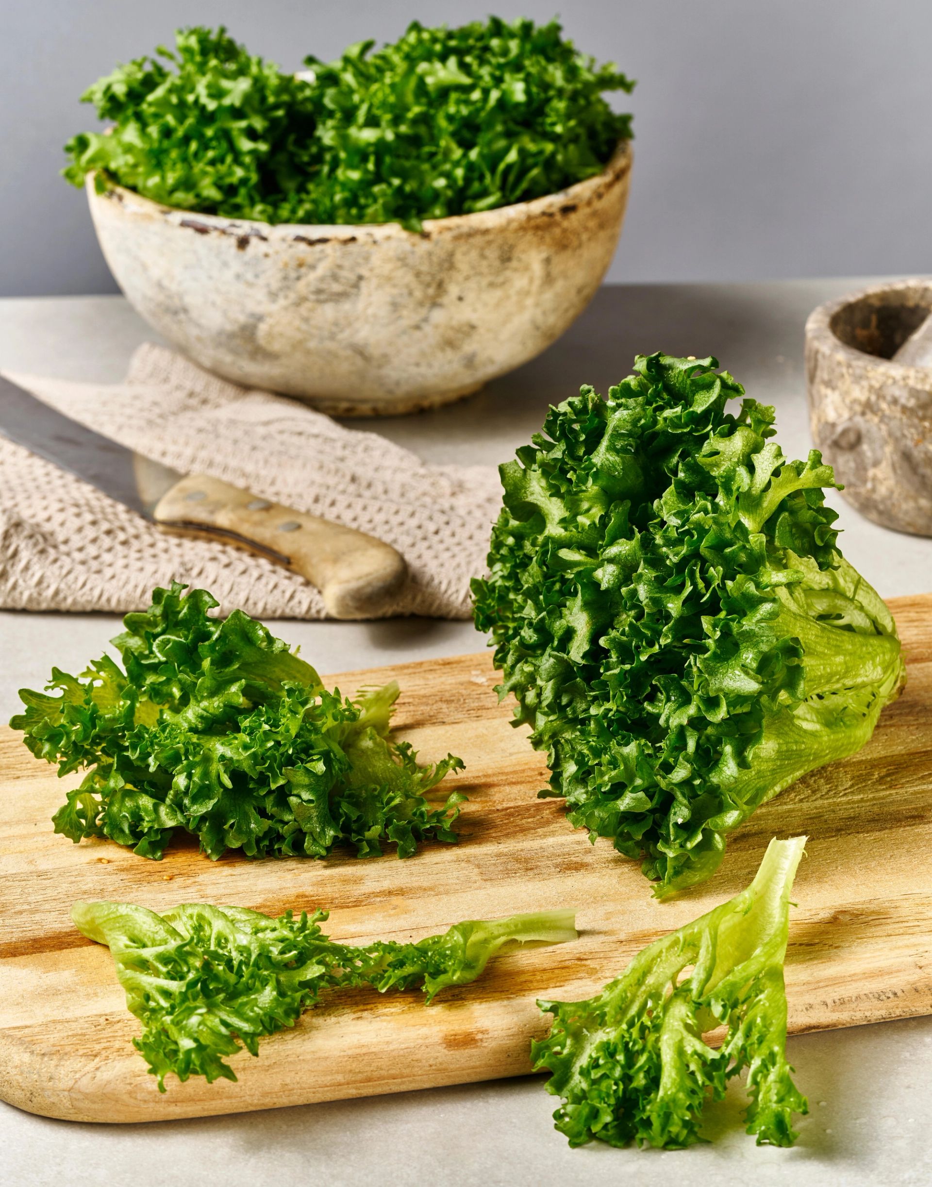 Grønn crispisalat salat ligger på et skjærebrett, både som hele blader og hakkede biter, med en skål salat og kjøkkenredskaper i bakgrunnen.