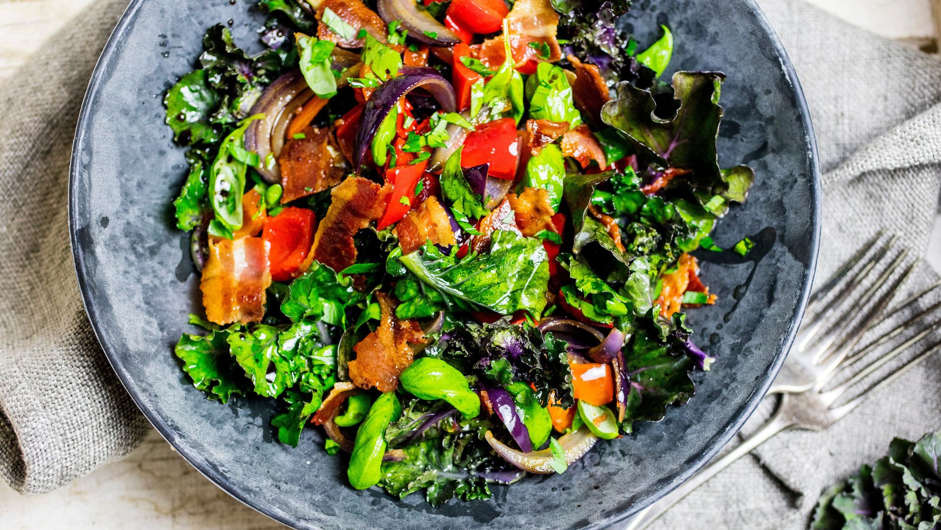 Fargerik salat servert i en stor, mørk skål. Salaten består av rosettkål, rødløk, paprika og sprø bacon. Ingrediensene er blandet med en blank dressing og toppet med frisk, hakket persille. Skålen står på et lyst underlag med en gaffel ved siden av.