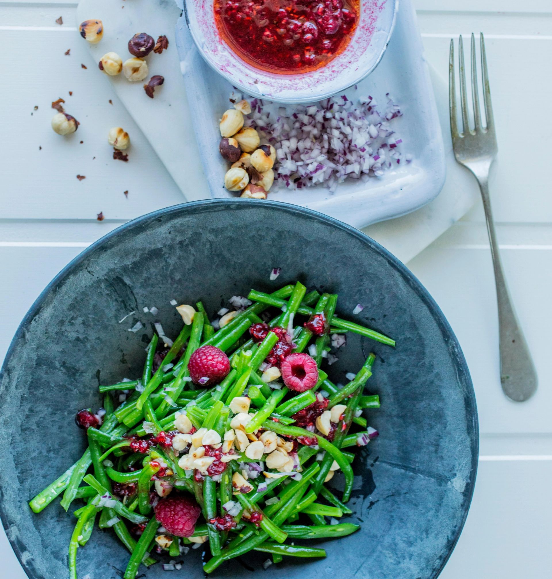 En mørk grå bolle fylt med en grønn bønnesalat toppet med bringebær og hakkede hasselnøtter. Ved siden av står et lite skjærebrett med en skål rød dressing, hakket løk og noen nøtter. En gaffel ligger øverst i bildet på en lys bordflate.
