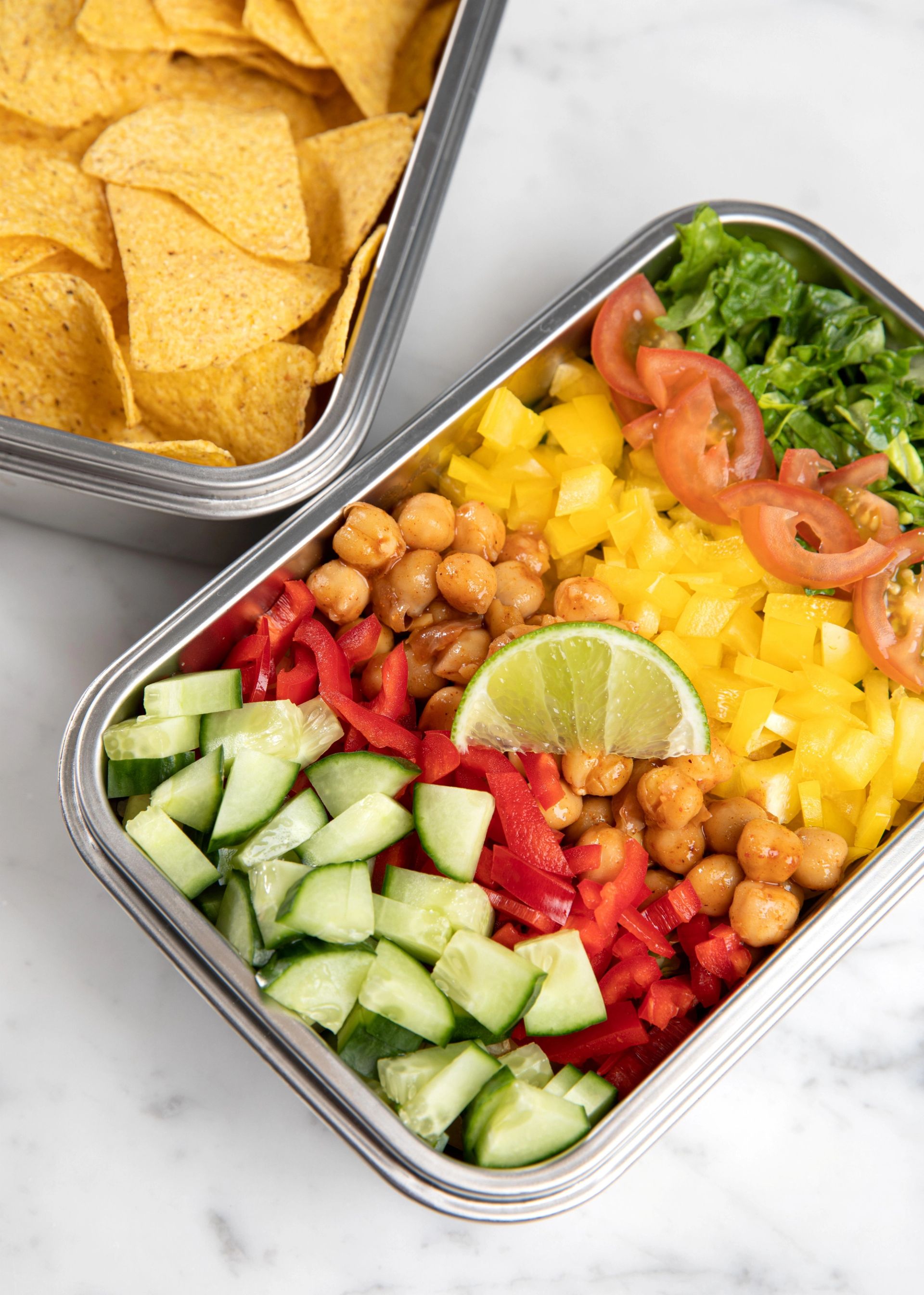 Matboks med fargerik salat av kikerter, gul og rød paprika, agurk, tomat og salatblader toppet med en limebåt, ved siden av en boks med tortillachips