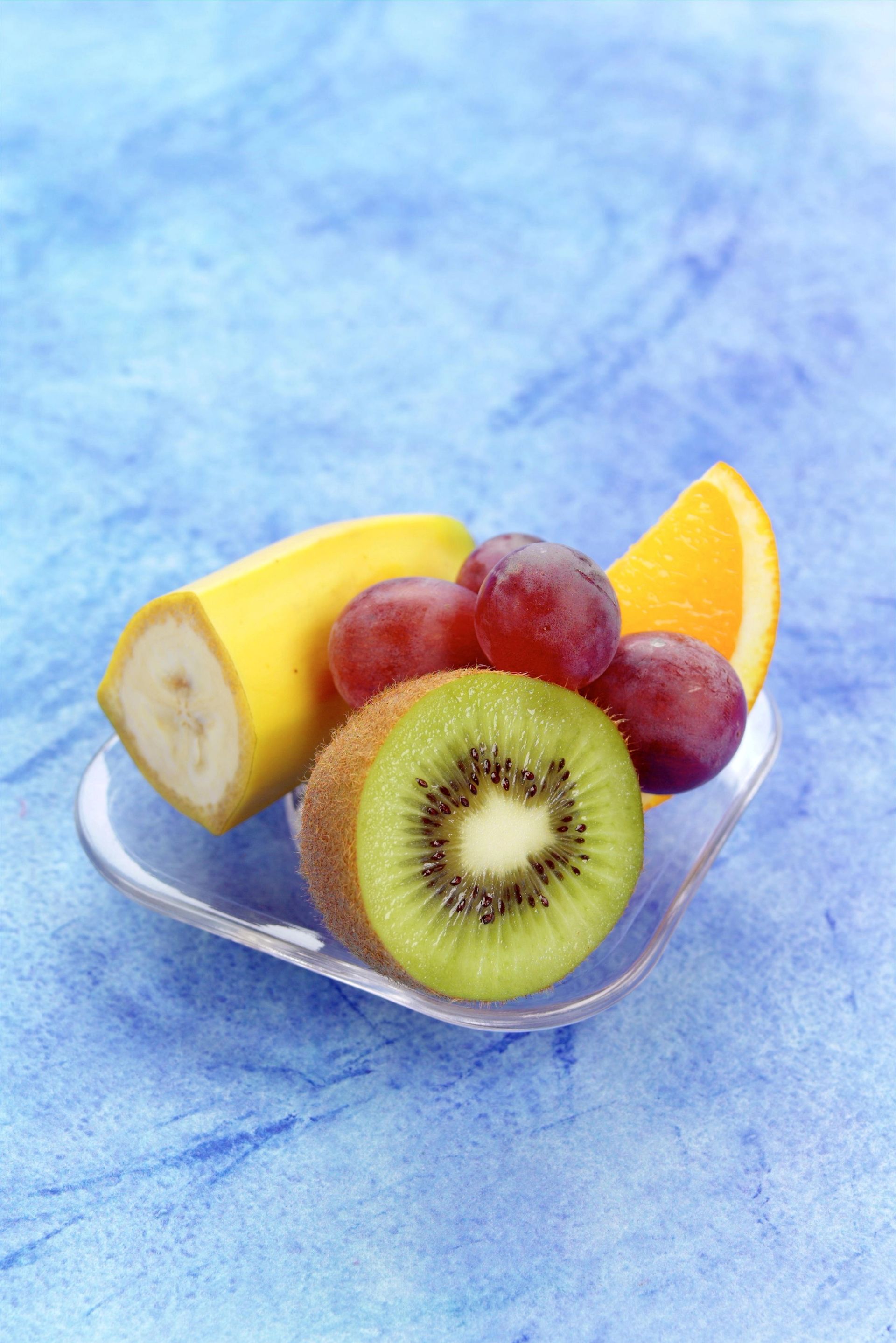 Glasskål med skiver av kiwi, druer, banan og appelsin plassert på blå bakgrunn.