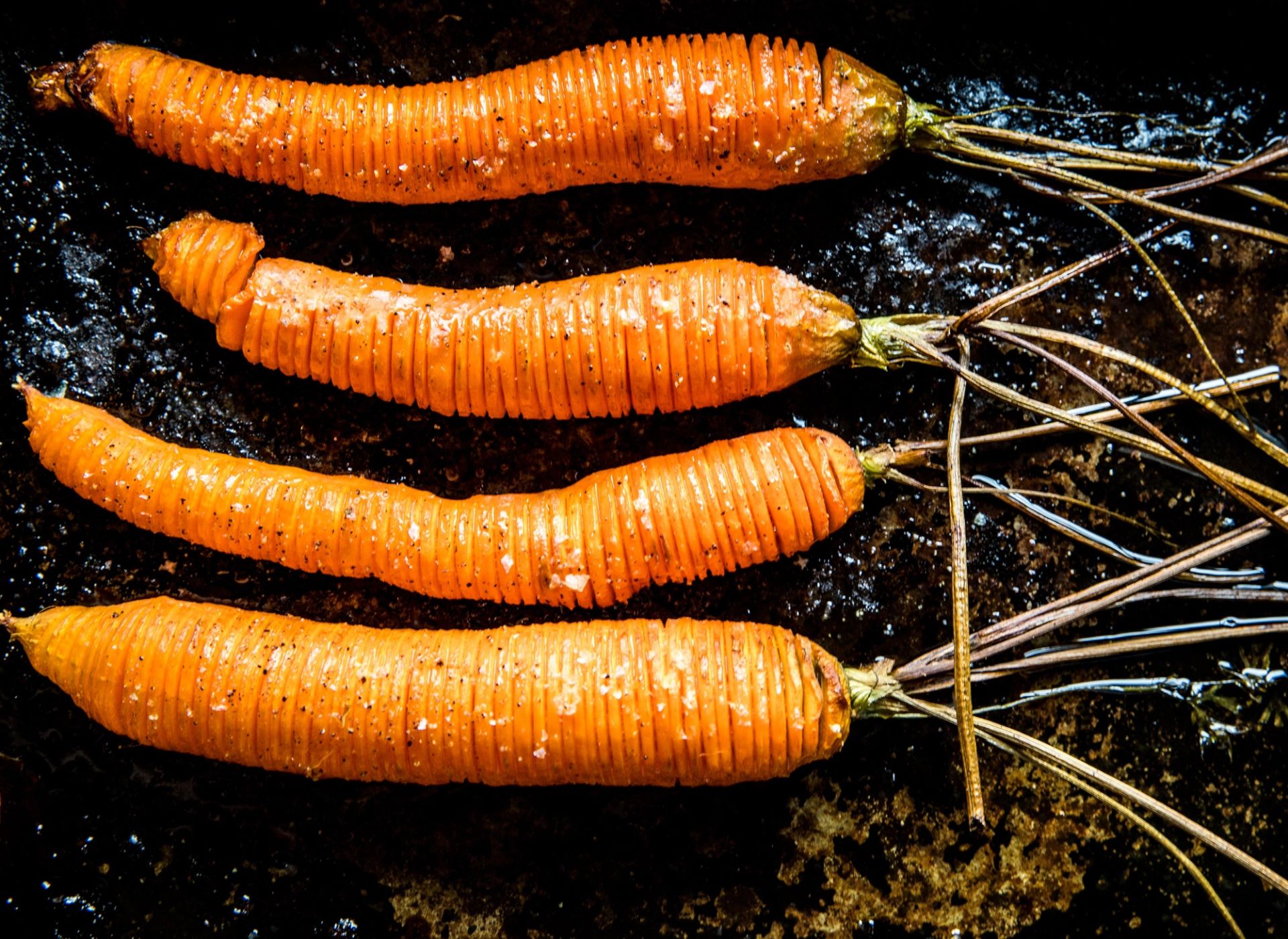 Fire ovnsbakte gulrøtter tilberedt som "hasselback" på et mørkt stekebrett. Gulrøttene har tette snitt på tvers, er penslet med olje og drysset med salt og pepper.