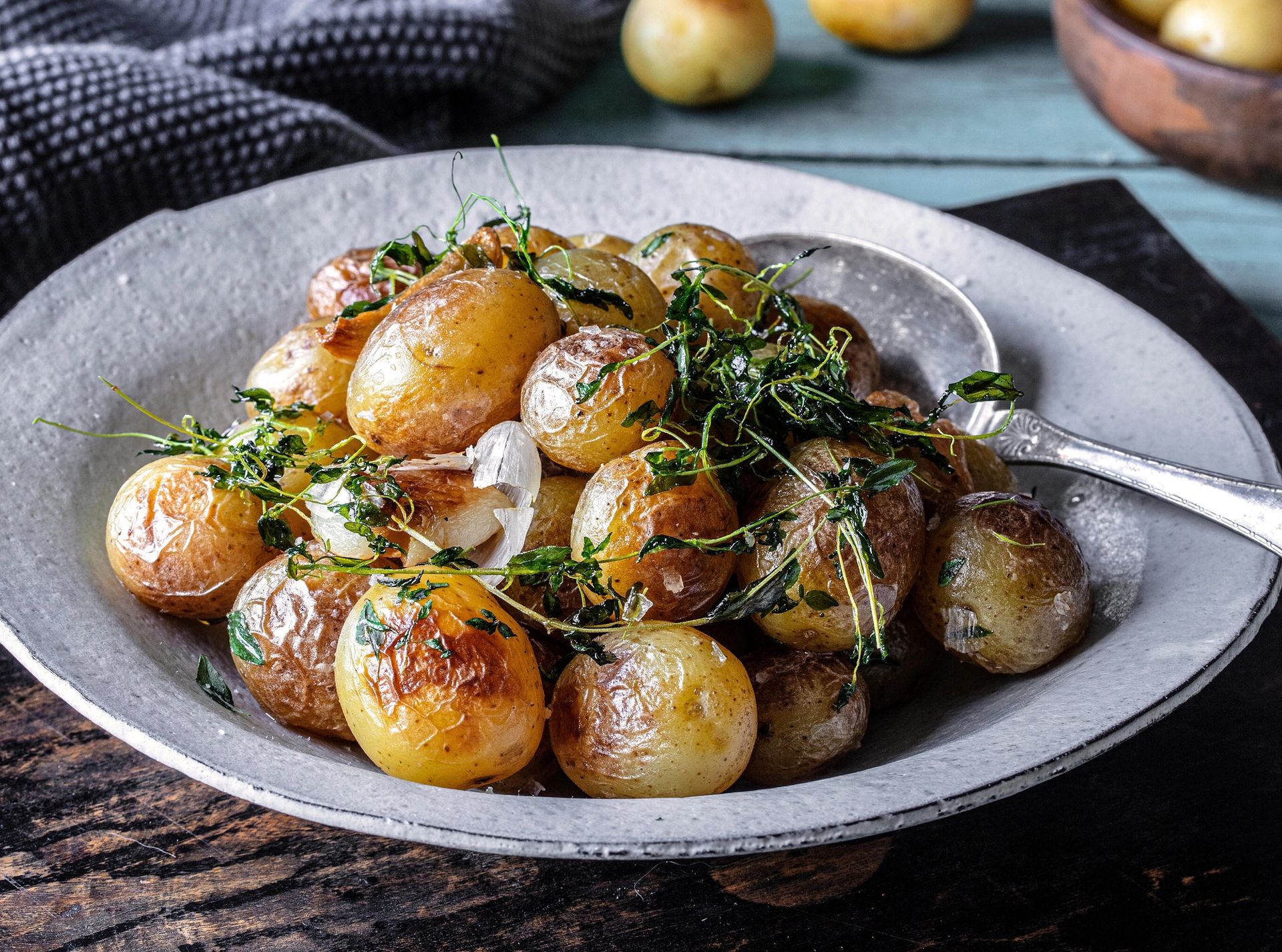 Små ovnsstekte mandelpoteter med gyllen og sprø overflate er servert i en lys skål og toppet med friske urter og løk. Potetene har fått en karamellisert stekeskorpe og fremstår som et smakfullt tilbehør til middag eller høytidsmåltid.