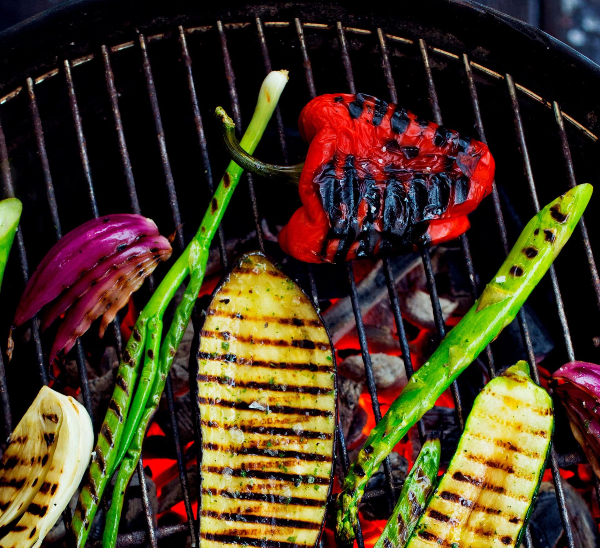 Grillede grønnsaker ligger på en grillrist over glødende kull. På risten ligger blant annet skiver av squash med grillstriper, asparges, en halv rød paprika med svidde kanter, og biter av rødløk.