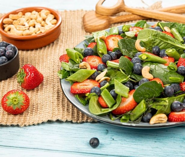 En fargerik sommersalat servert på en grå tallerken. Salaten inneholder frisk baby leaf mix, skivede jordbær, blåbær, sukkererter og cashewnøtter.
