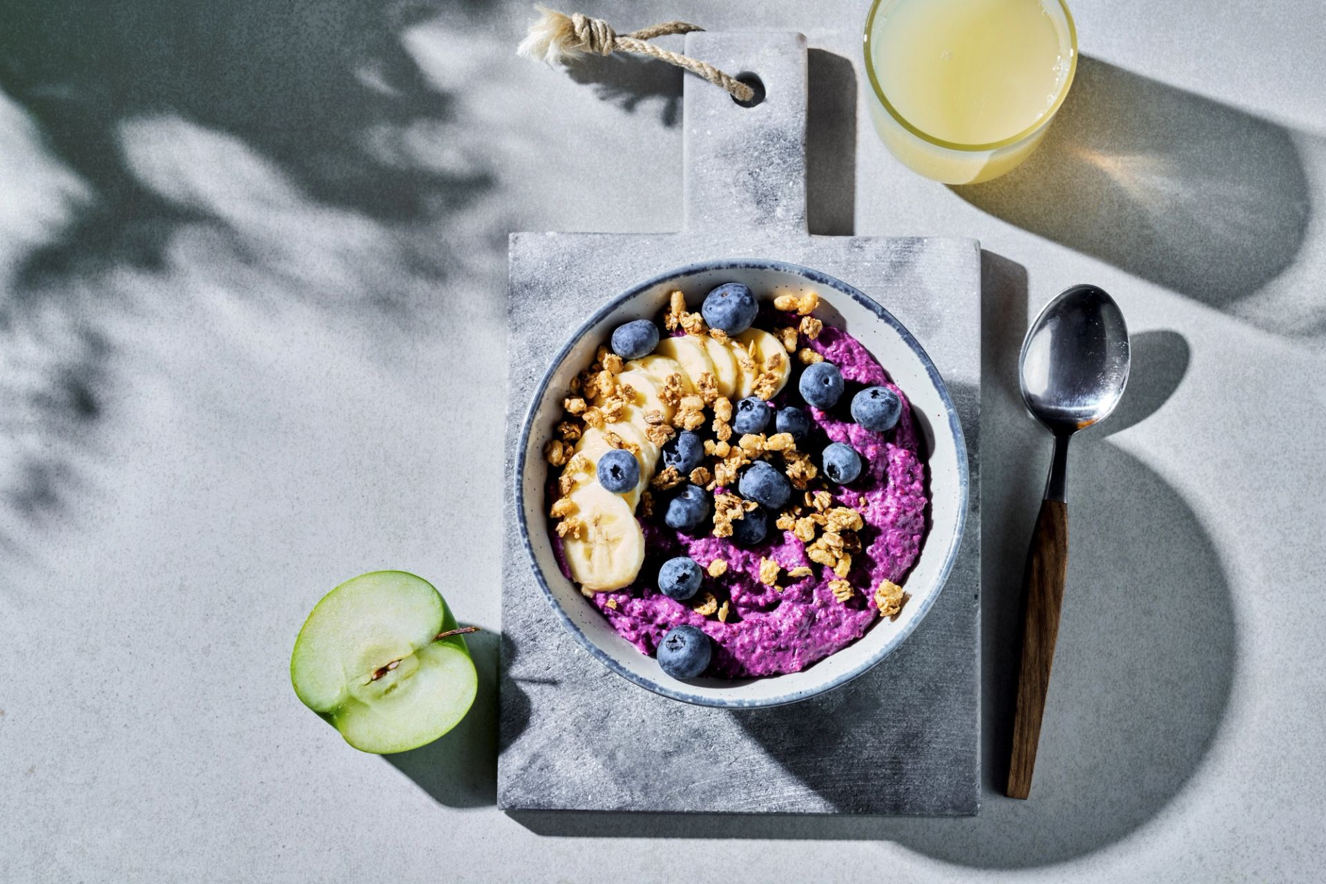 Smoothiegrøt i en skål med lilla bærbase, toppet med blåbær, skiver av banan og granola. Skålen står på et lite steinbrett, med skje, eple og et glass juice ved siden av.