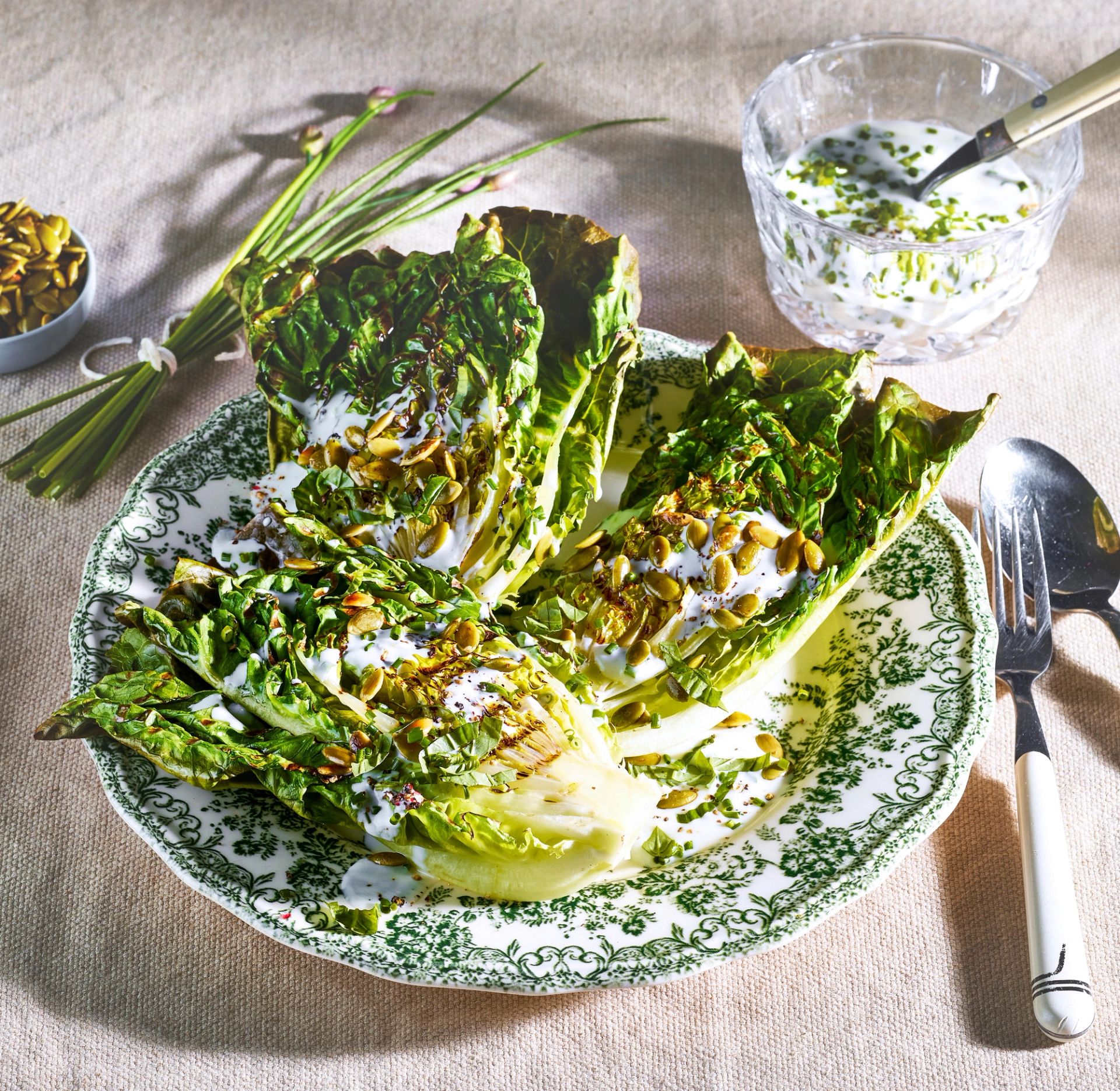 Grillede hjertesalathalvdeler servert på et mønstret fat, toppet med ristede frø og en kremet dressing med urter. En skål med dressing og friske urter ligger i bakgrunnen.