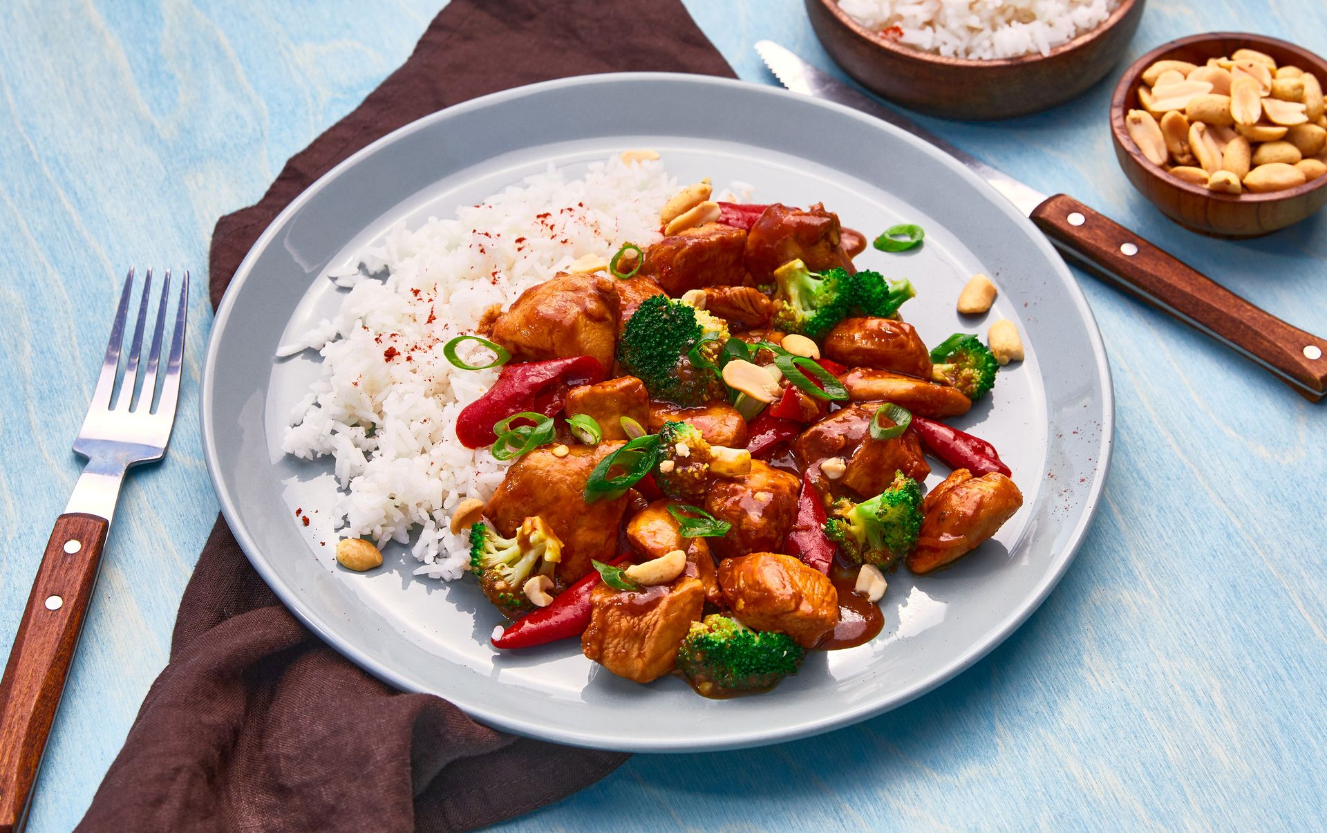 Stekt brokkoli Kung Pao servert med kyllingbiter, rød paprika og ris på en grå tallerken. Retten er toppet med knasende peanøtter og snittet vårløk. Ved siden av tallerkenen ligger bestikk med trehåndtak, samt små skåler med ekstra ris og peanøtter, alt plassert på et lyseblått tregulv.