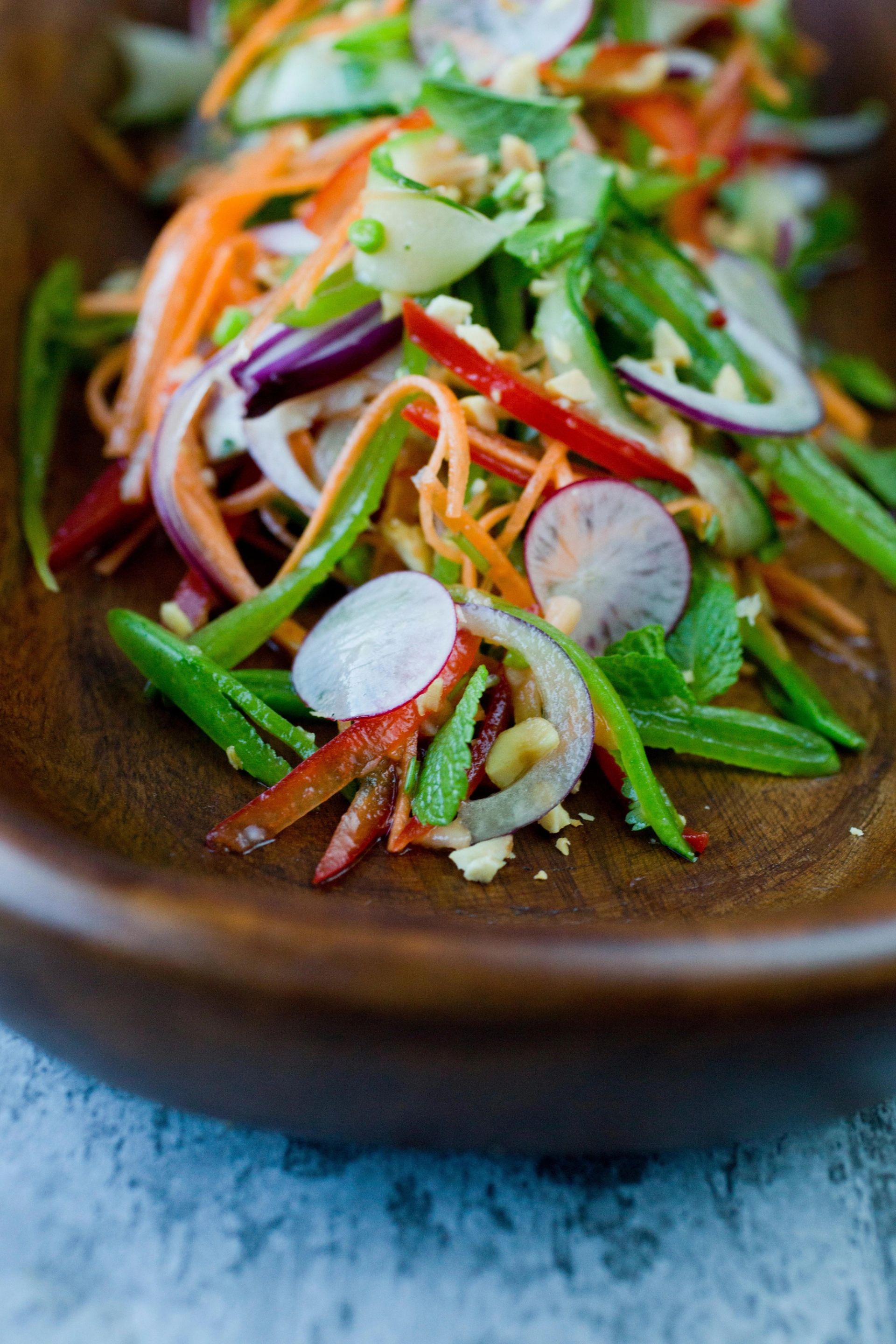 En fargerik råkostsalat med tynne strimler av gulrot, paprika, agurk, reddik og urter servert i en oval trebolle.