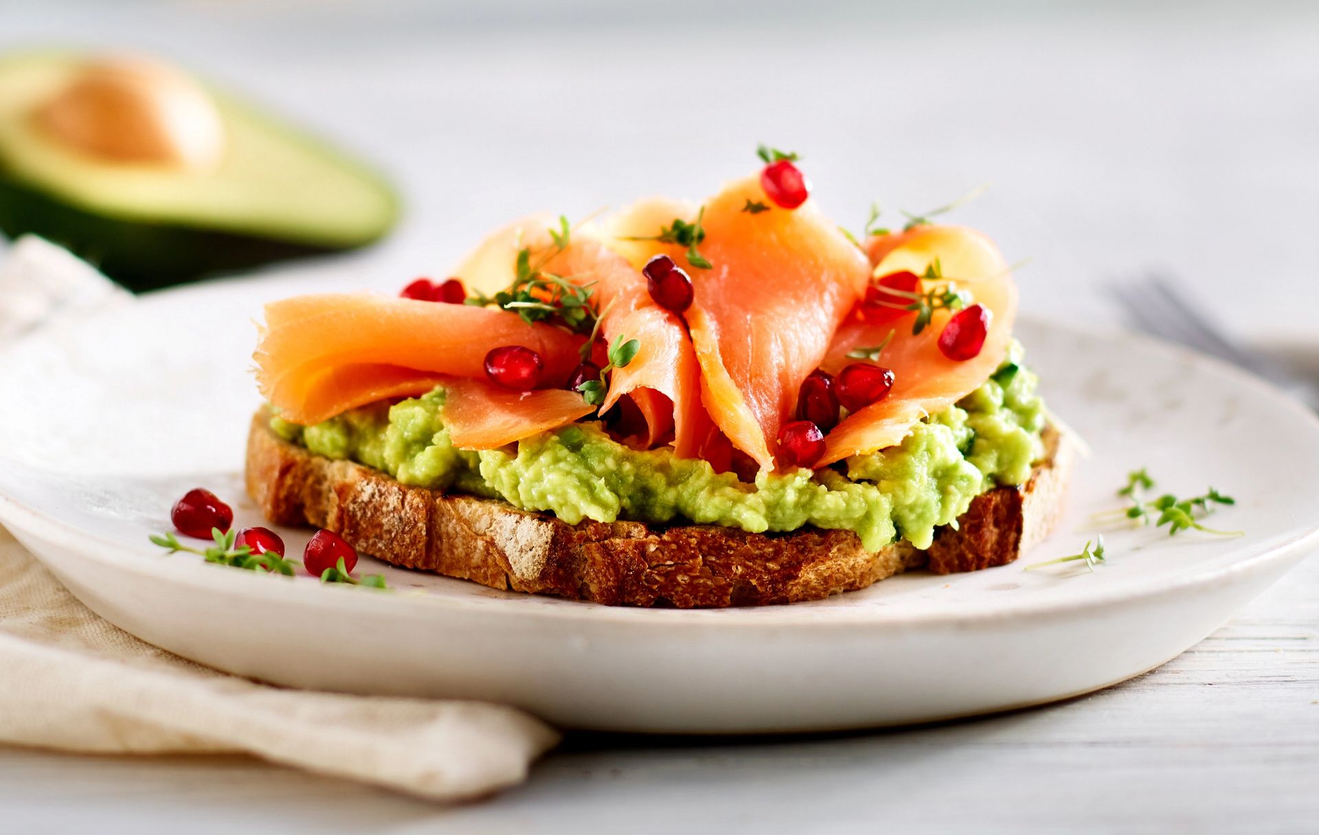 Ristet brød toppet med most avokado, silkemyke skiver røkelaks og granateplekjerner, garnert med friske urter og servert på en hvit tallerken, med en delt avokado i bakgrunnen.