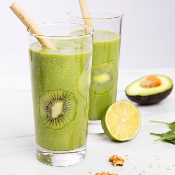 To høye glass fylt med grønn smoothie med hele skiver av kiwi presset mot innsiden av glassene. Begge glassene har lyse sugerør. Ved siden av ligger en halv lime, en delt avokado og noen valnøttbiter på en lys bordflate.
