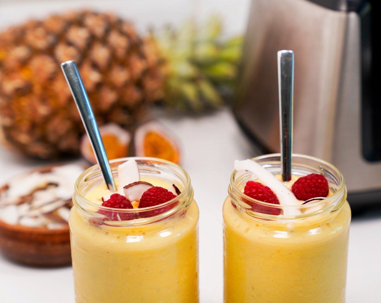 Gul og frisk ananassmoothie i glass, toppet med bringebær og kokosflak, med fersk ananas i bakgrunnen.