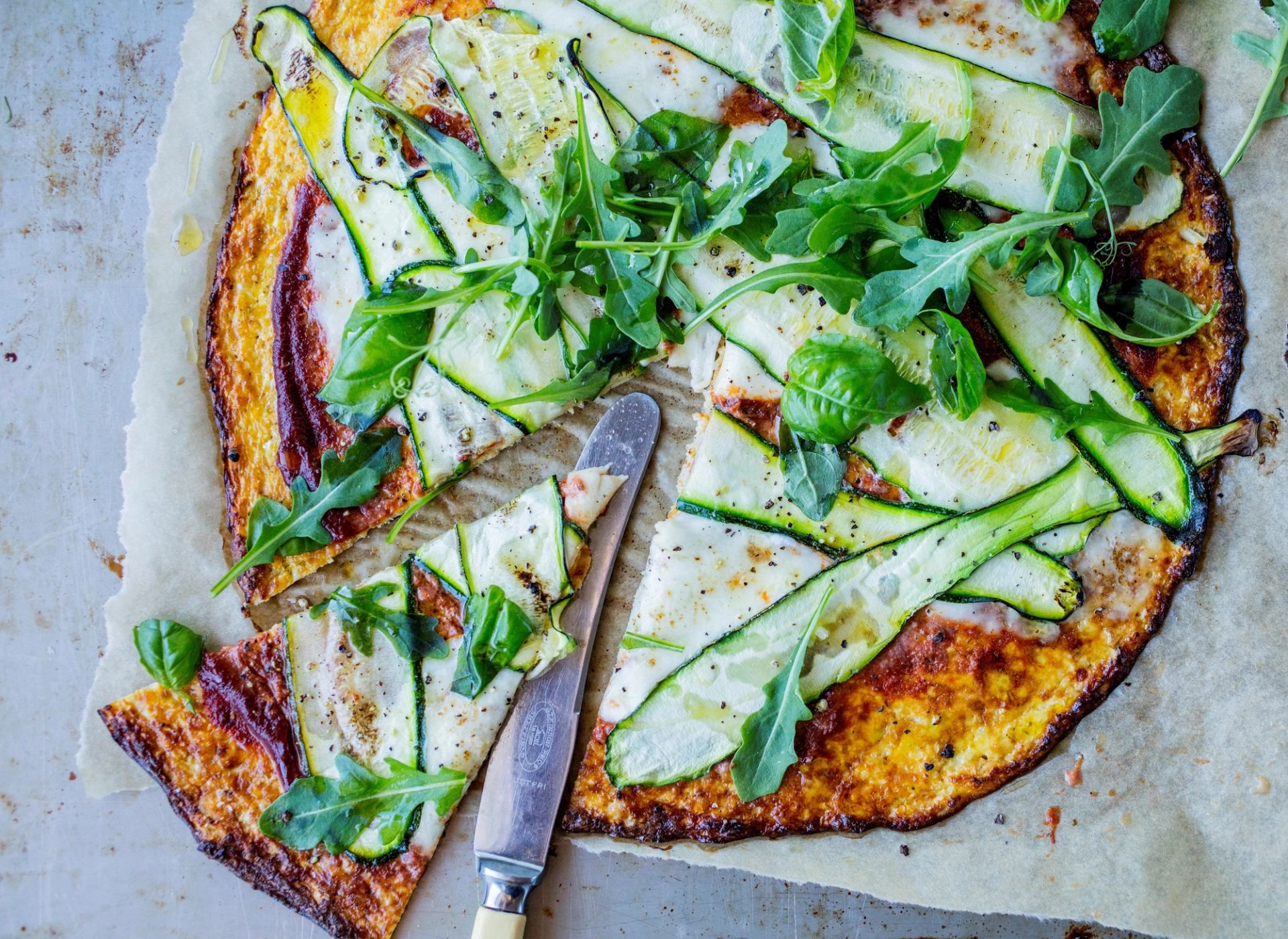 En gyllen og sprø blomkålpizza på bakepapir, toppet med tomatsaus, smeltet mozzarella og tynne skiver squash. Pizzaen er garnert med frisk ruccola og basilikum, og et stykke er skåret ut med en kakekniv ved siden av.