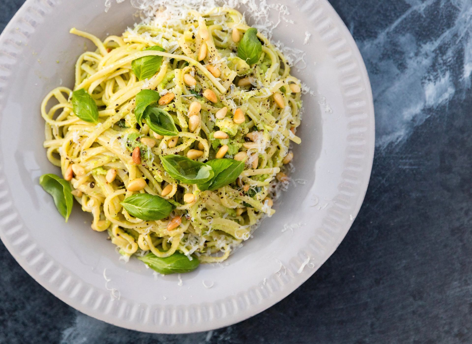 Tallerken med pasta vendt i avokadopesto, toppet med pinjekjerner, fersk basilikum og revet parmesan på mørk bakgrunn.