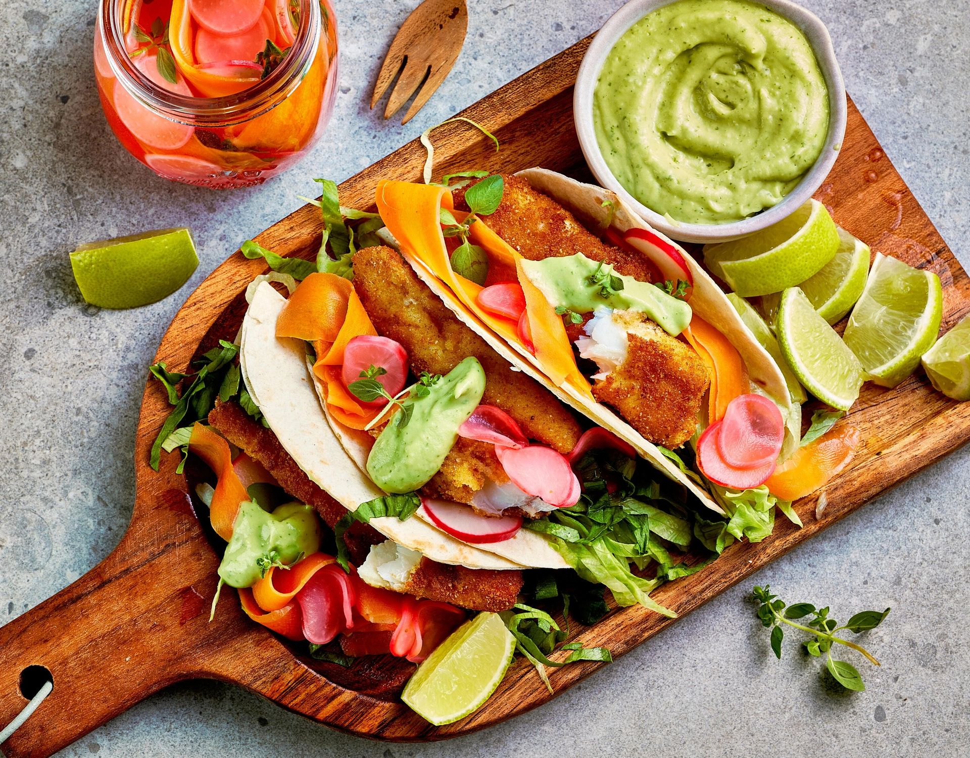 To tacos med sprø, panert fisk servert i myke tortillalefser med ruccola, syltede reddiker og kremet grønn avokadosaus på et rustikt serveringsbrett. Rundt tacoene ligger båter av lime, og i bakgrunnen står et glass med syltede grønnsaker.
