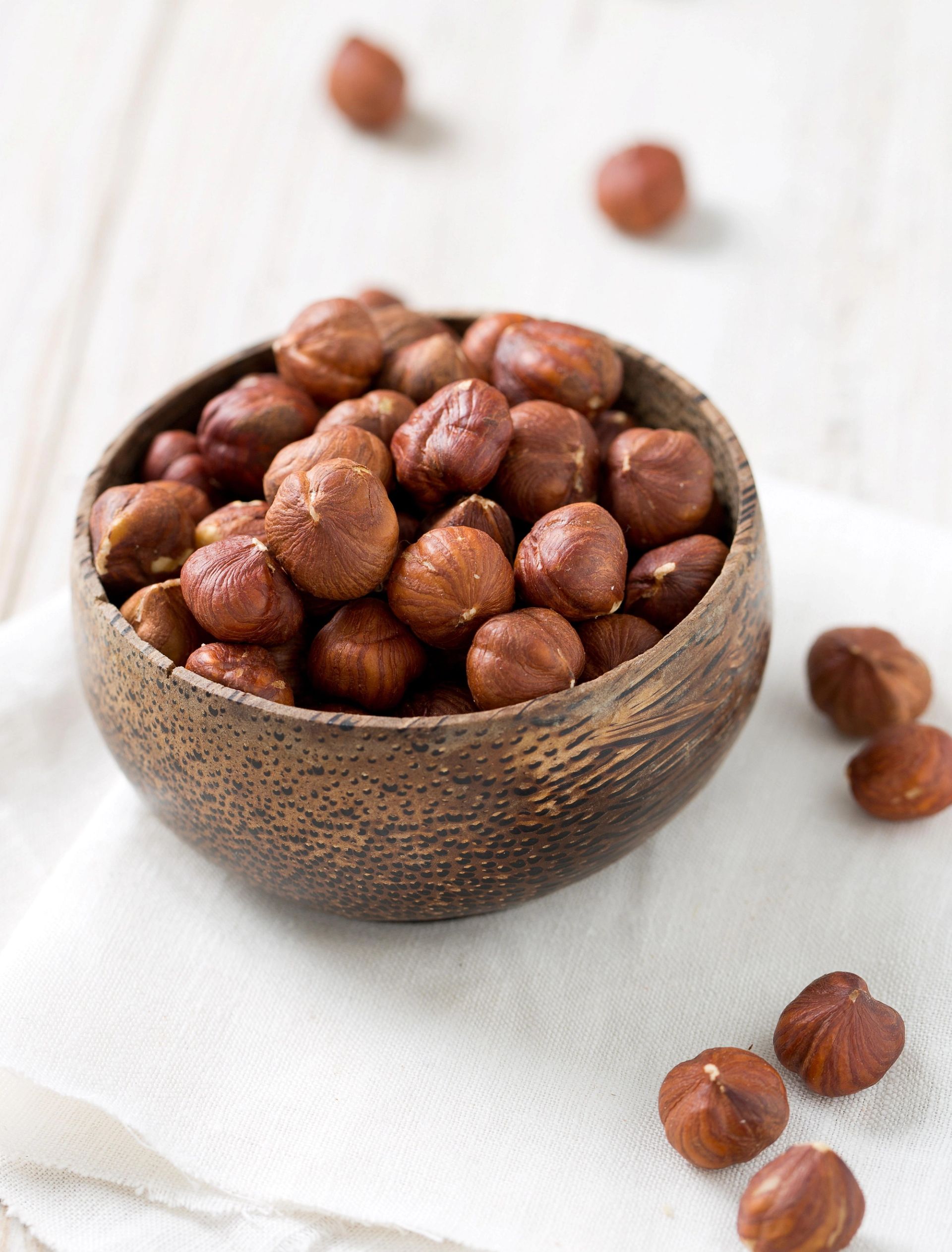 En liten, rund bolle med rustikk overflate er fylt med hasselnøtter og står på et lyst underlag. Noen få hasselnøtter ligger spredt rundt bollen på bordet, og bakgrunnen er myk og nøytral uten tydelige elementer.