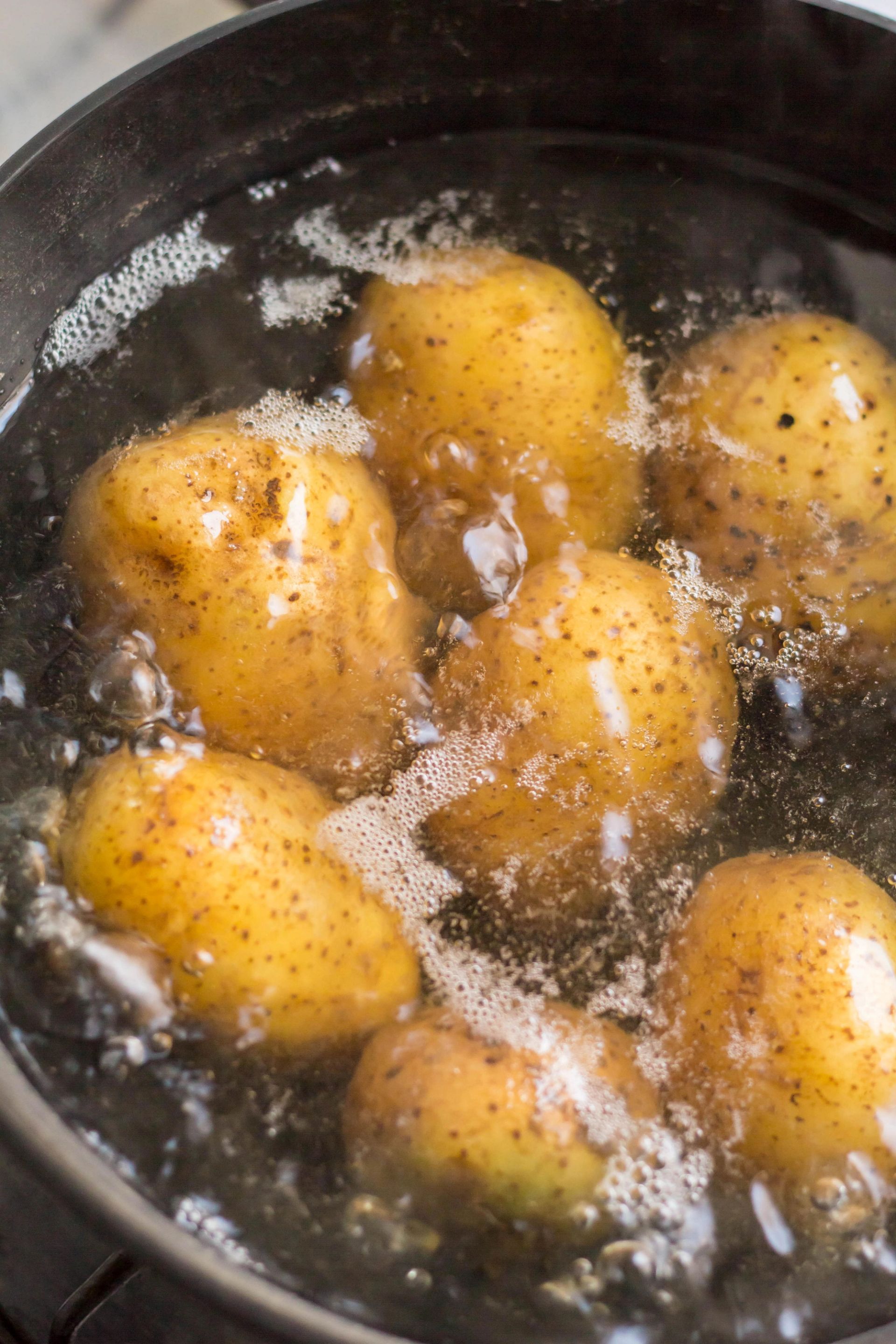 Poteter koker i fossende vann i en kjele, med bobler og skum på overflaten og potetskall synlig gjennom vannet.