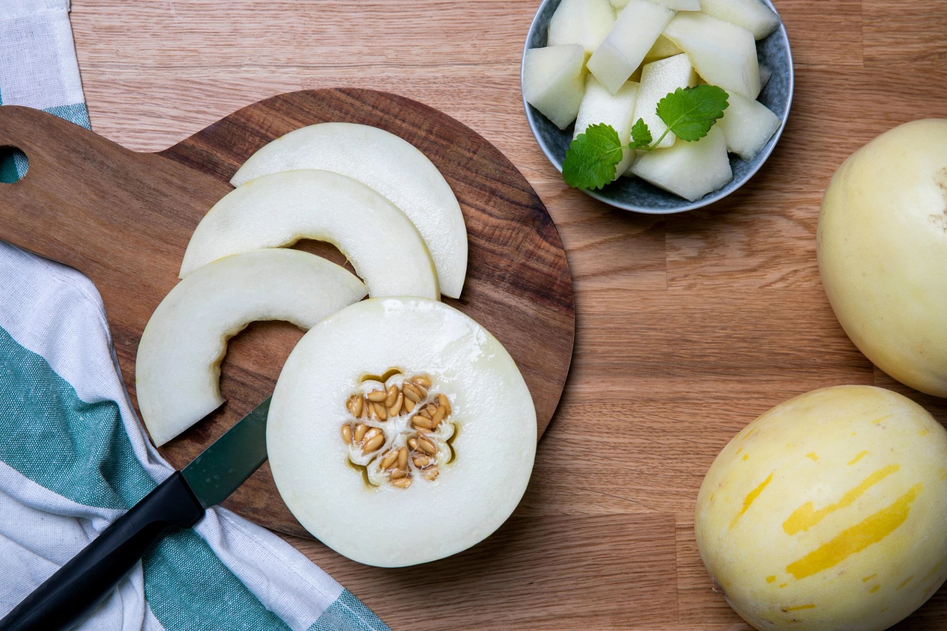 Skivet snowball‑melon på et rundt skjærebrett med kniv, frø synlige i midten. En skål med melonbiter og hele snowball‑meloner på et trebord