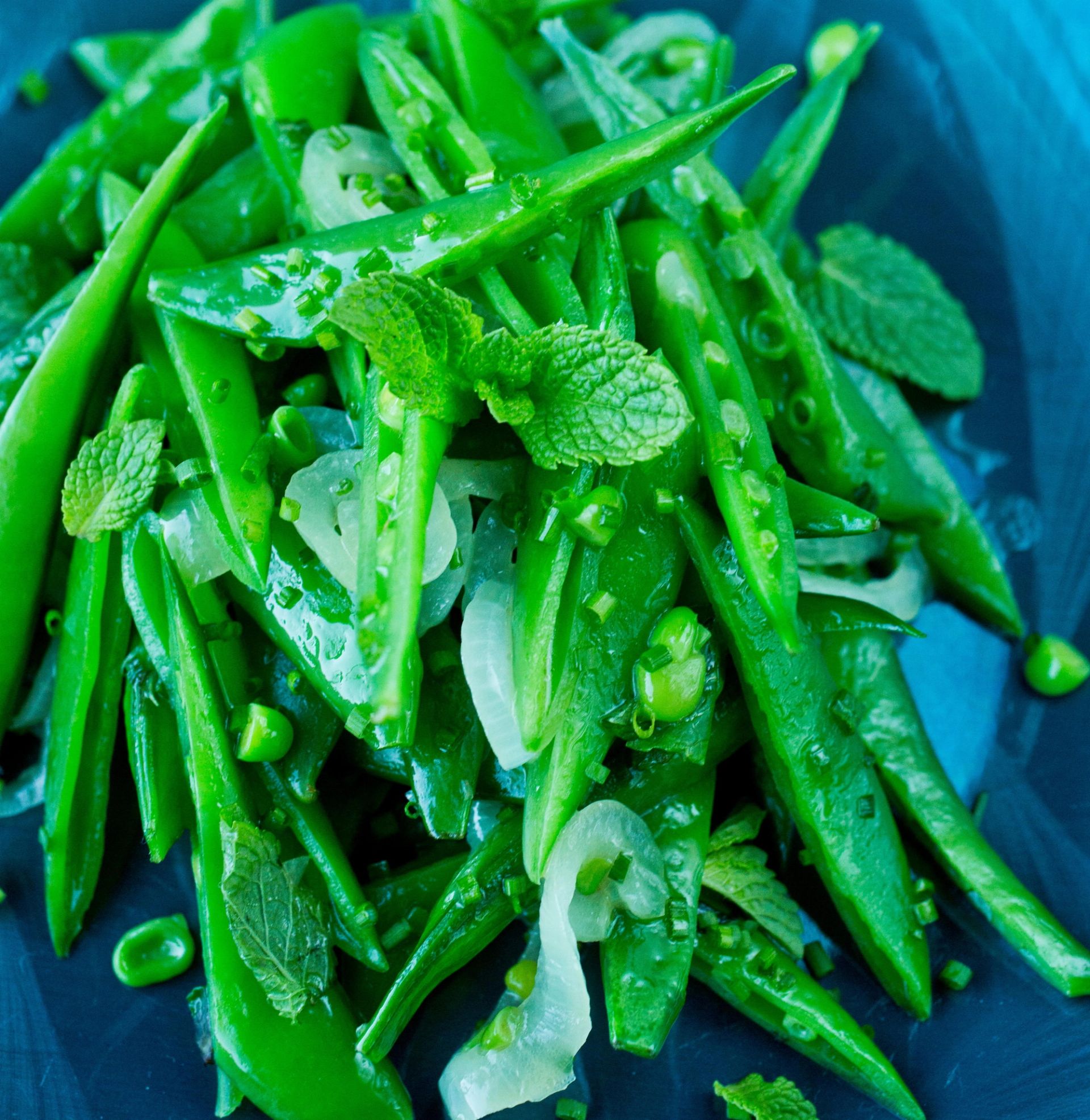 Nærbilde av en frisk grønn salat med sukkererter og grønne bønner, blandet med tynne skiver løk, erter og friske urter som mynte, servert i en blå bolle.