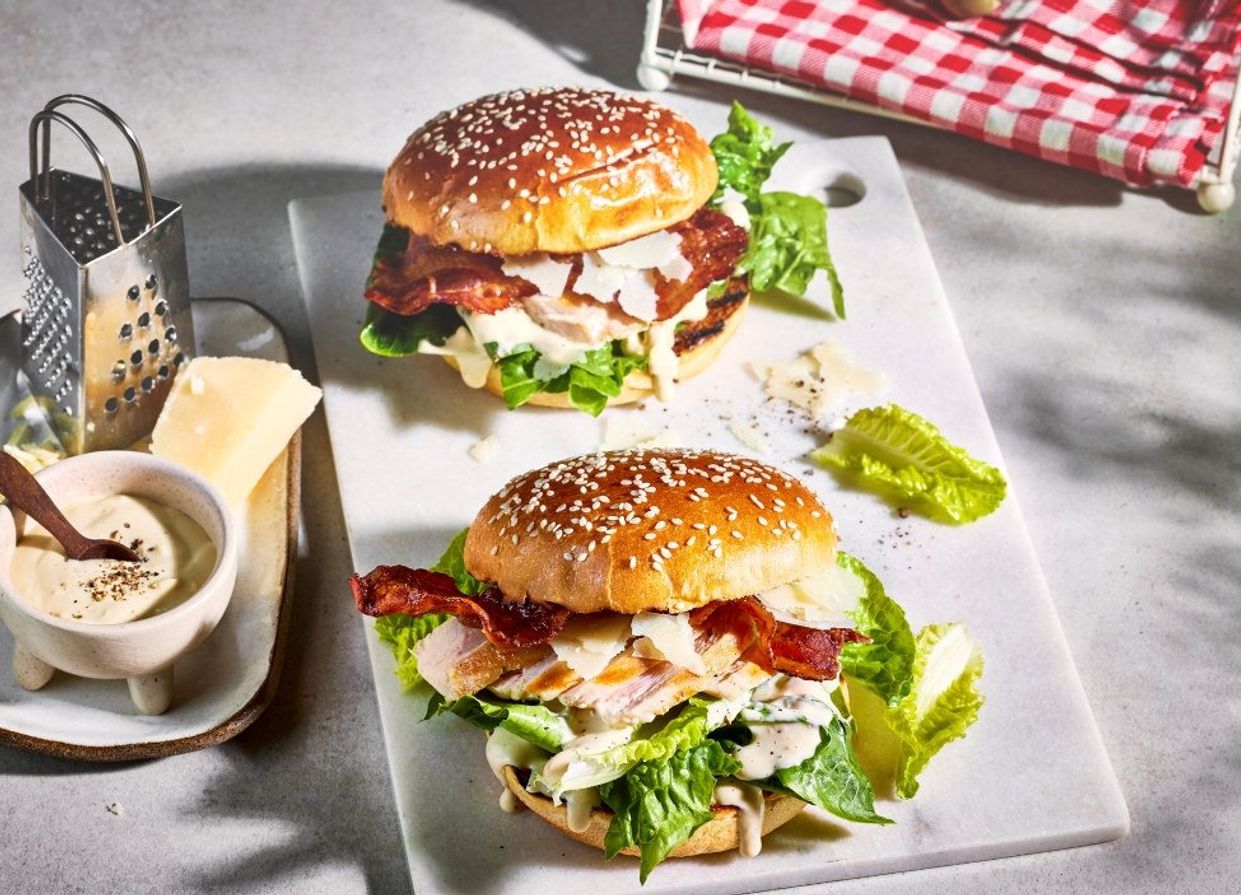 To burgere med sesambrød ligger på et lyst fat, fylt med salat, kyllingfilet, sprøstekt bacon og dressing, servert med revet ost og en liten rasp ved siden av.