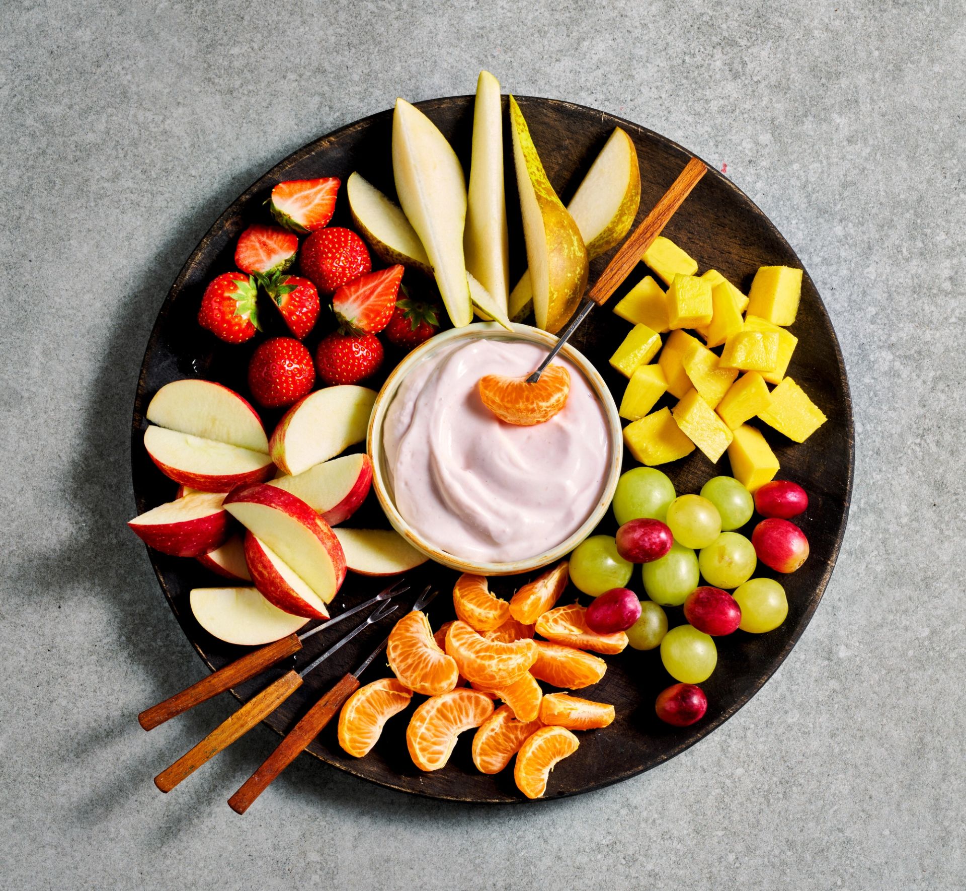 Fruktfat med oppskåret frukt som eple, pære, jordbær, druer, appelsin og mango arrangert rundt en skål med rosa yoghurtdipp. Fatet er sett ovenfra og inneholder også små serveringsgafler, noe som gjør det enkelt å dele som mellommåltid eller servering til flere.