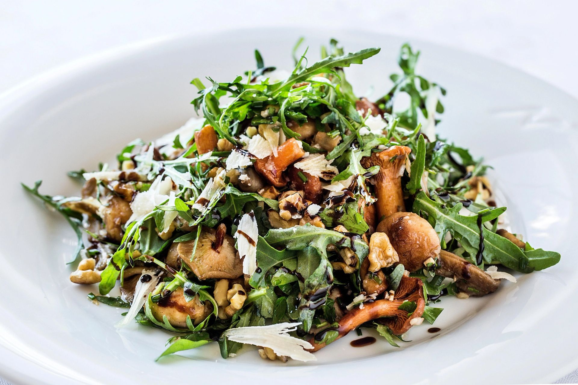 Salat med ruccola, stekte sopper, hakkede nøtter og revet ost, toppet med en mørk dressing, servert på en hvit tallerken