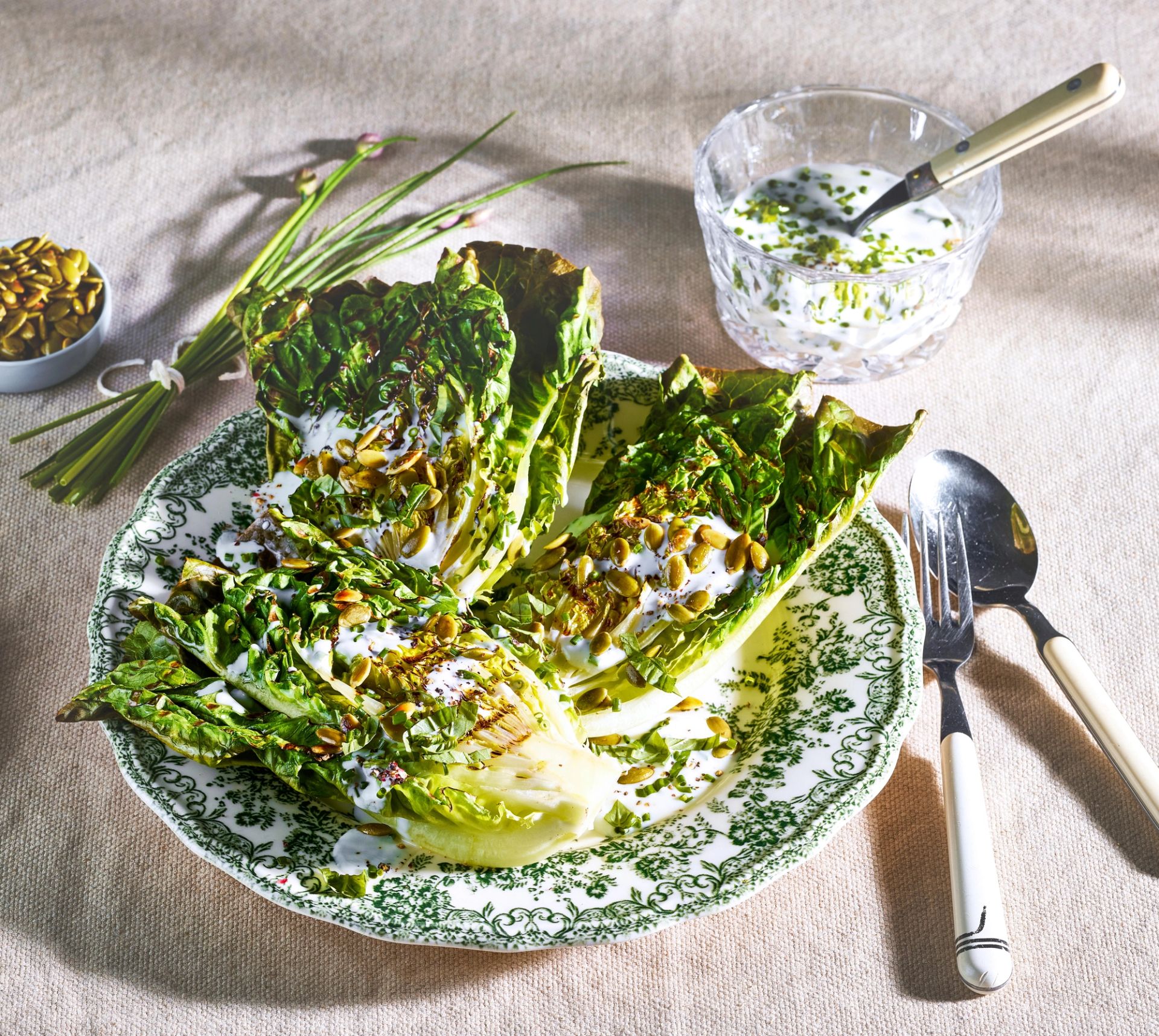 Grillet hjertesalat delt i båter ligger på et mønstret fat, toppet med kremet dressing, ristede frø og urter, med bestikk og en skål dressing plassert rundt på et lyst kjøkkenbord.