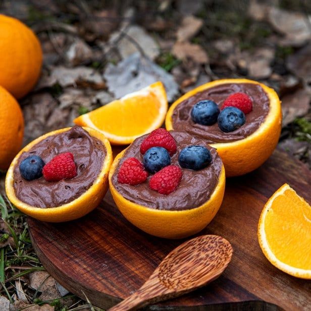 Halve appelsiner fylt med sjokolademousse, toppet med bringebær og blåbær, servert på et trebord utendørs med friske appelsinbåter ved siden av.