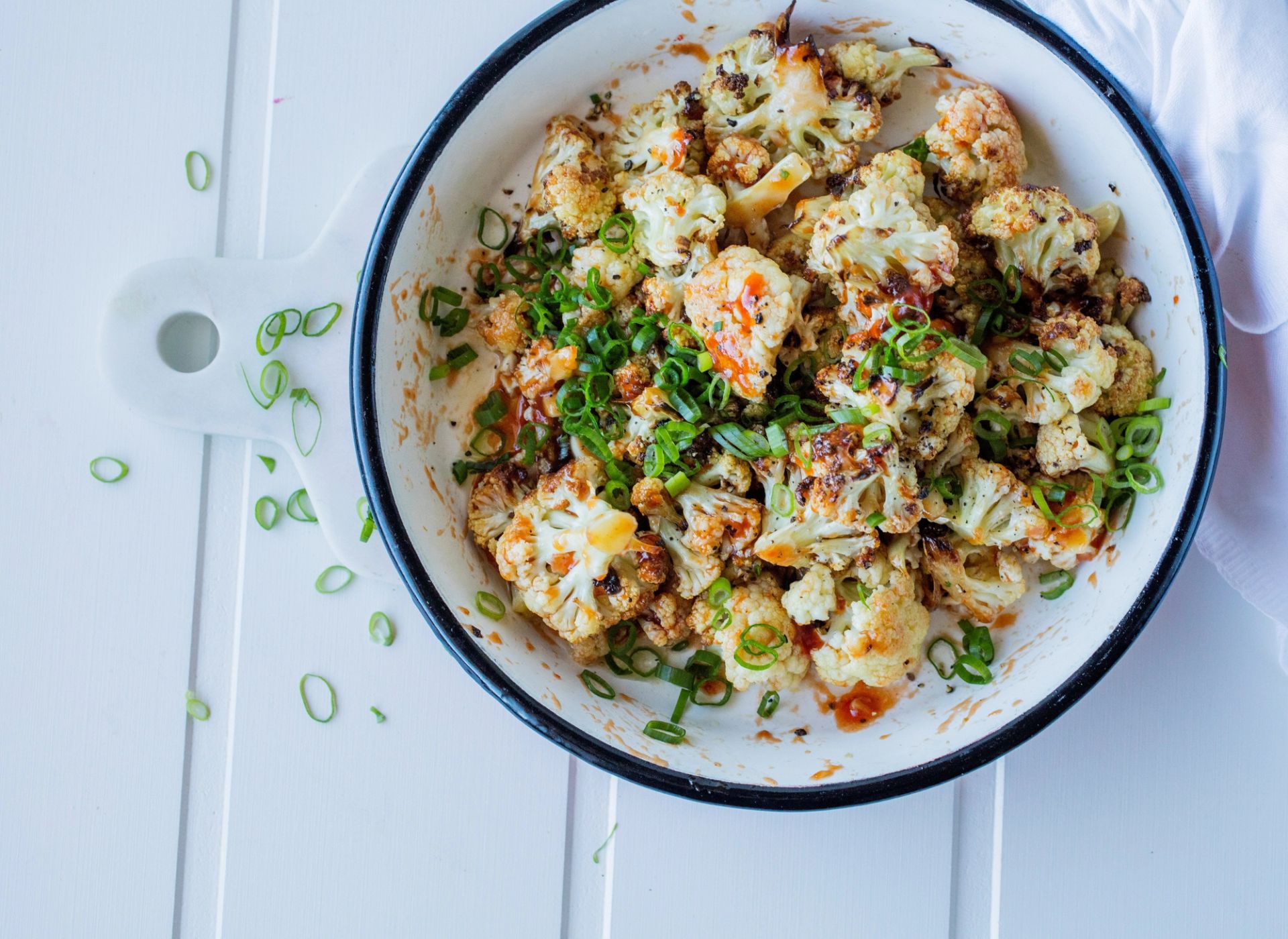 Ovnsbakt blomkål i en hvit, emaljert panne, sett ovenfra. Blomkålbukettene er gylne og lett svidde i kantene, og er toppet med chilisaus og rikelig med finsnittet vårløk.