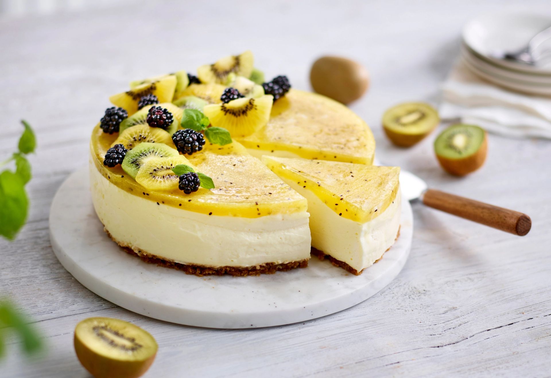 Ostekake med sprø kjeksbunn og lokk av sitrongelé og gul kiwi, toppet med bjørnebær, kiwi og mynte, servert på et rundt kakefat.