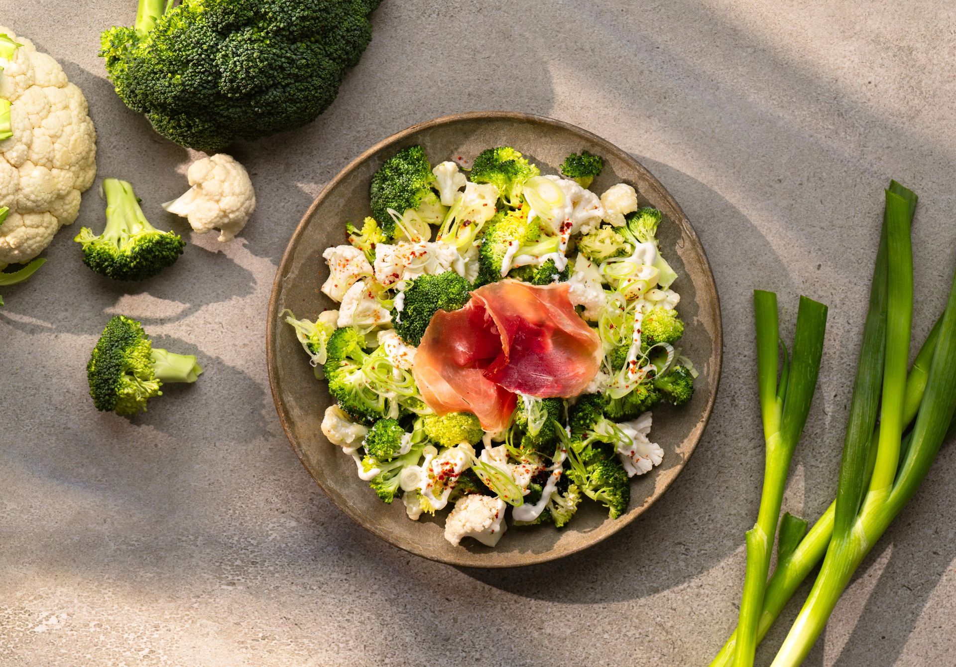 Lun salat med bakt brokkoli og blomkål, fetaostkrem, vårløk og spekeskinke servert på et fat.